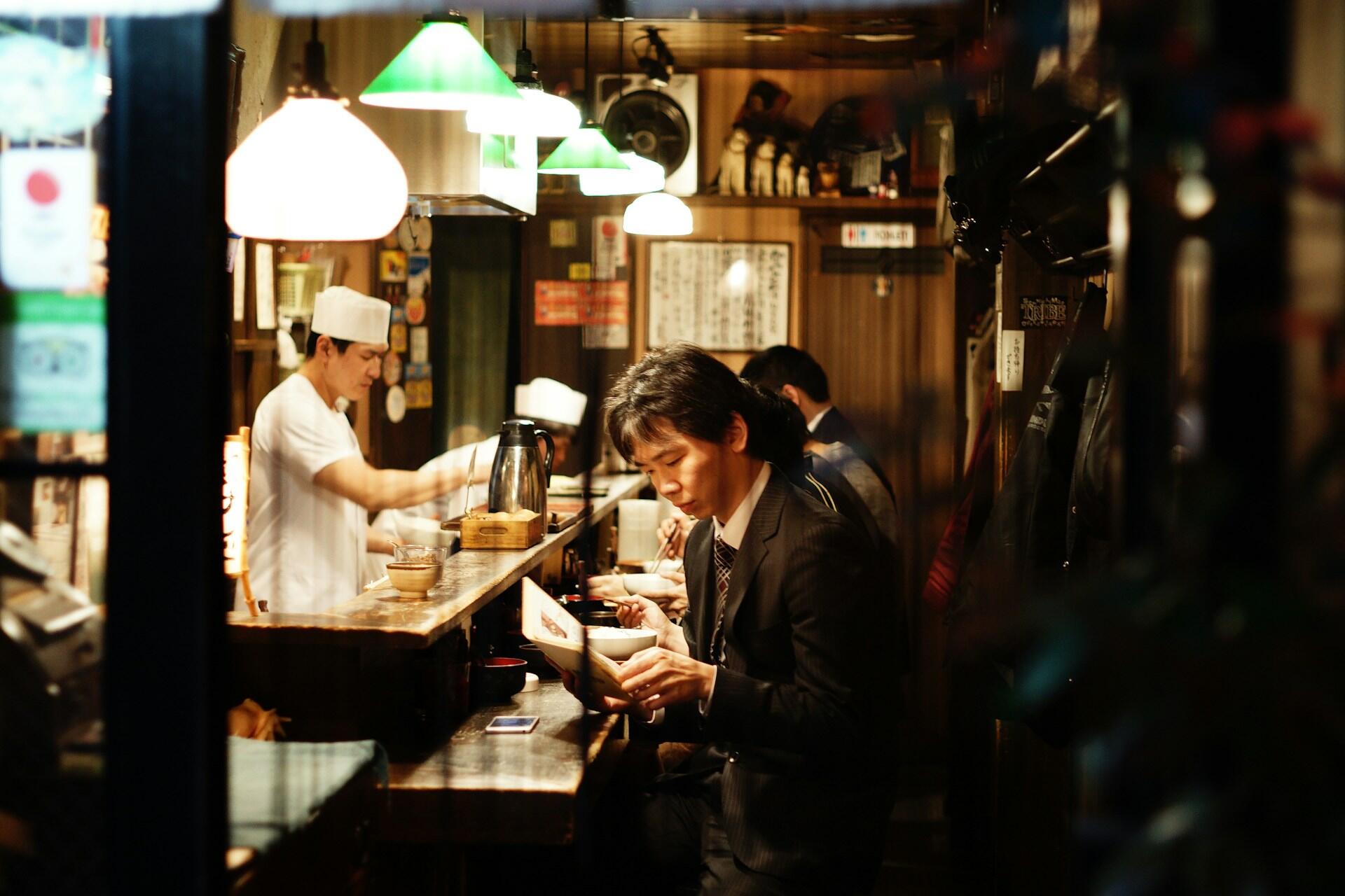 A cozy restaurant interior with hanging green lamps, featuring a chef at the counter and patrons enjoying their meals.