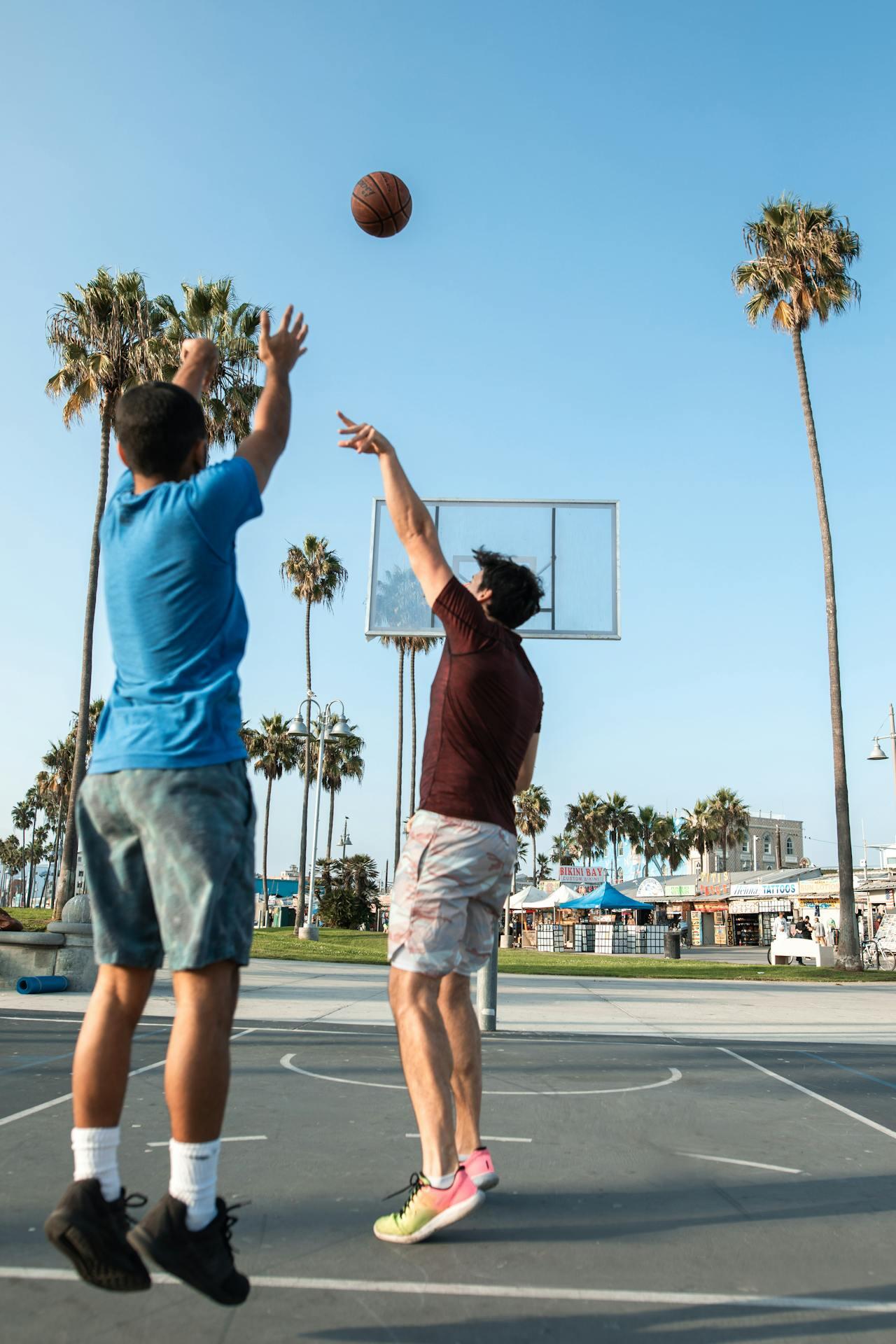 Two young men playing basketball outdoors, one shooting while the other prepares to catch the ball, with palm trees in the background.