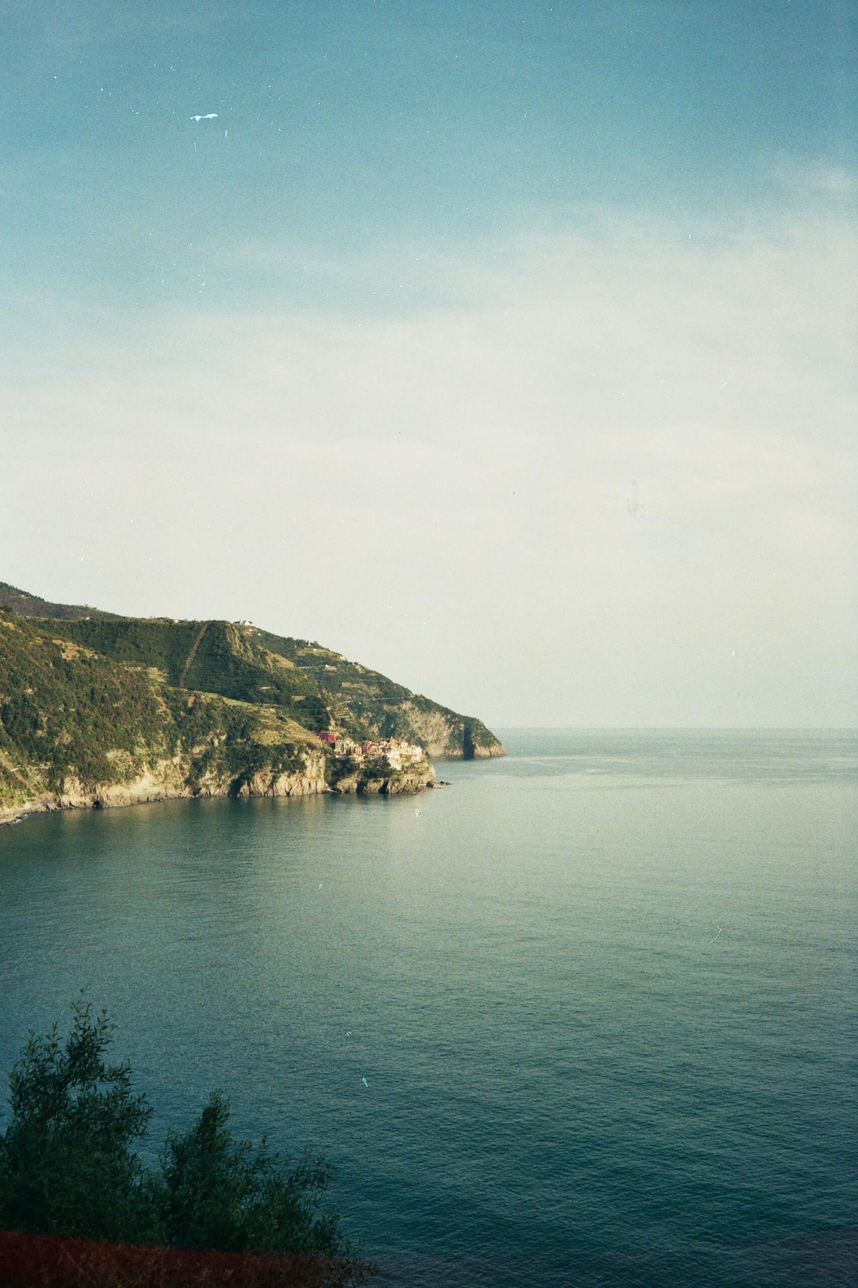 Tranquil coastal view featuring lush green cliffs descending into calm blue waters under a soft sky, with a village dotting the shoreline.