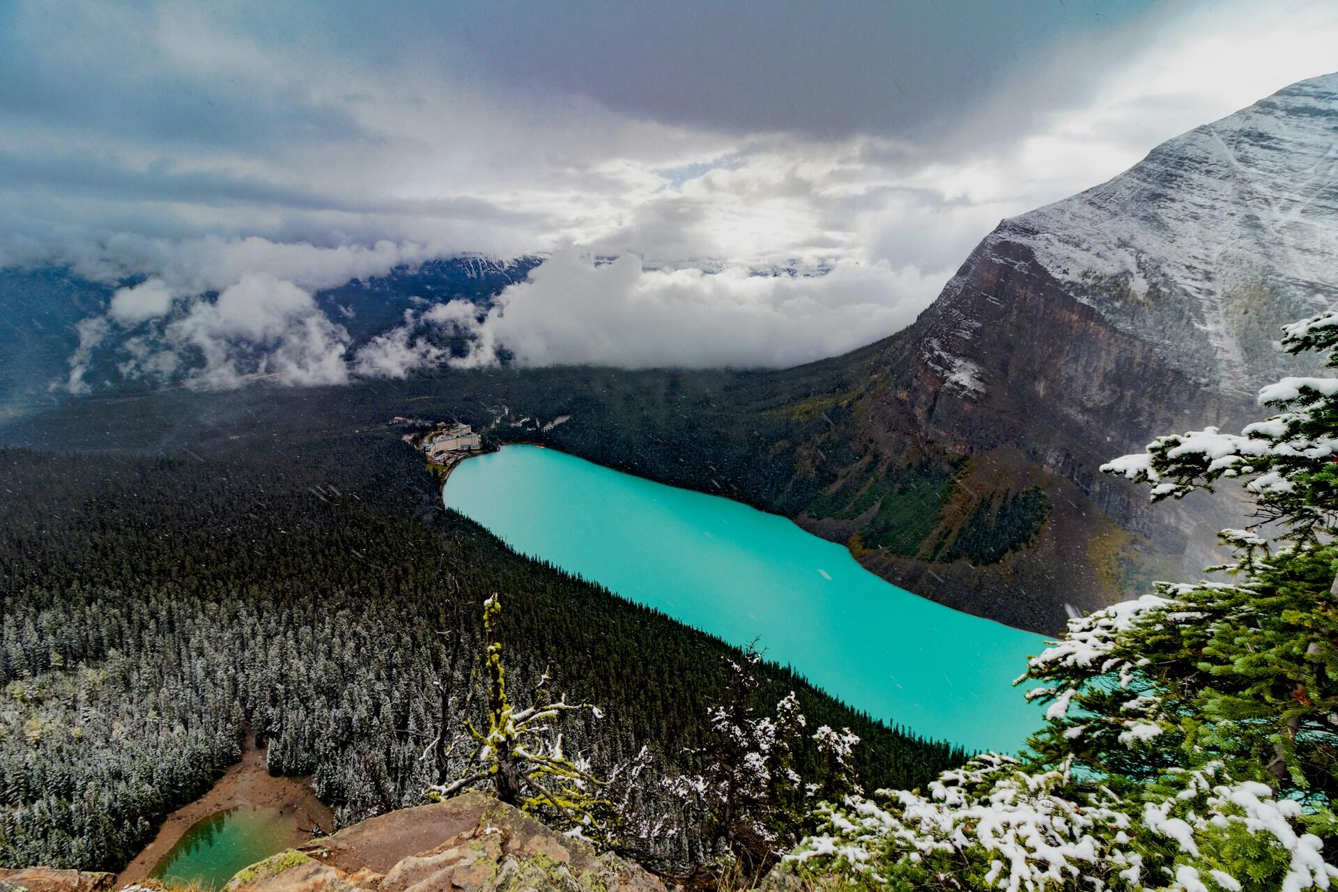A breathtaking aerial view of a turquoise lake surrounded by dense evergreen forests and rugged mountains under a cloudy sky.