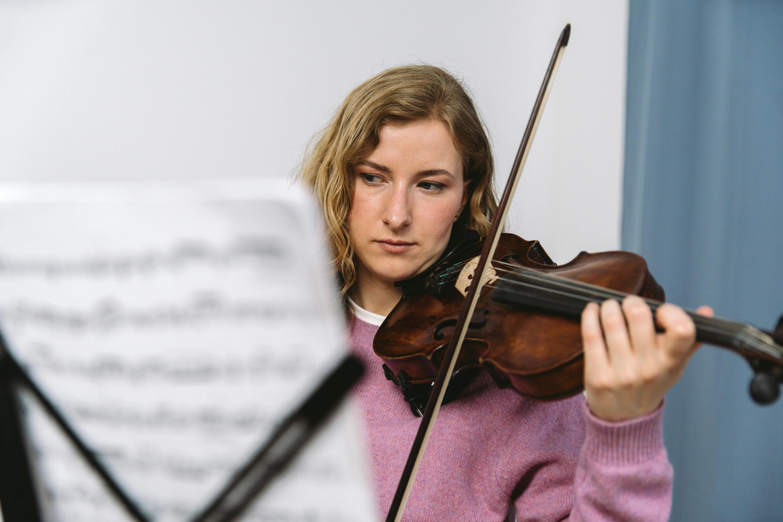 A woman in a pink sweater plays the violin in front of sheet music.