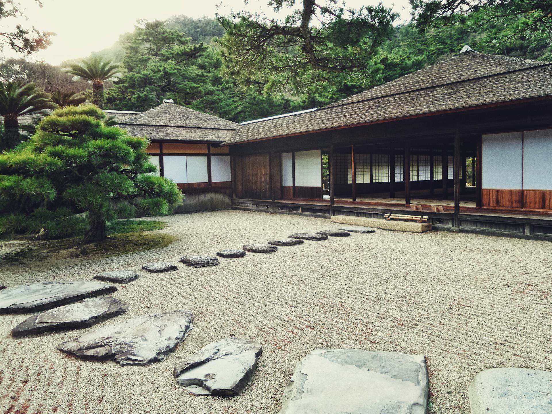 A serene Japanese garden featuring stone paths, a lush pine tree, and traditional wooden buildings amidst a tranquil landscape.
