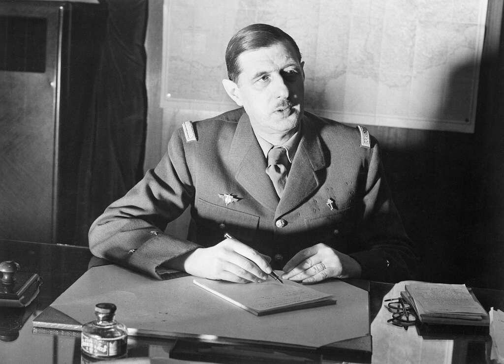 A man in a military uniform sits at a desk, writing on a pad of paper, with maps visible in the background.
