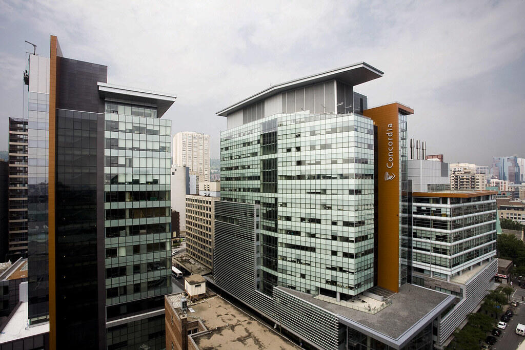 A wide-angle view of Concordia University’s modern campus buildings in downtown Montreal. The structures are made of glass and steel, with one building prominently displaying the “Concordia” name on a tall orange vertical panel. Surrounding the campus are other high-rise buildings under a cloudy sky.