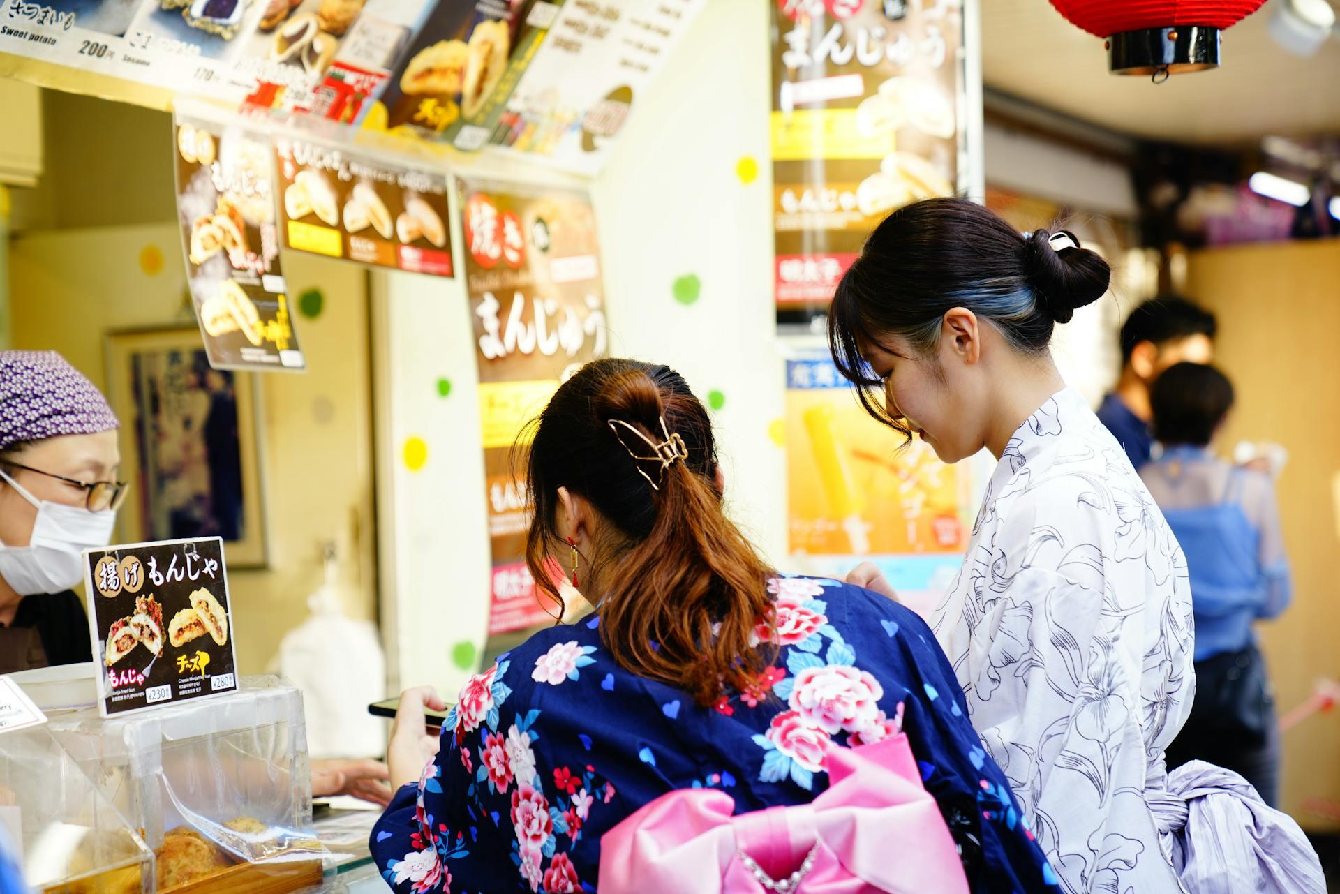 Two women in colorful traditional kimonos stand at a food stall, discussing options with a vendor, surrounded by vibrant posters.