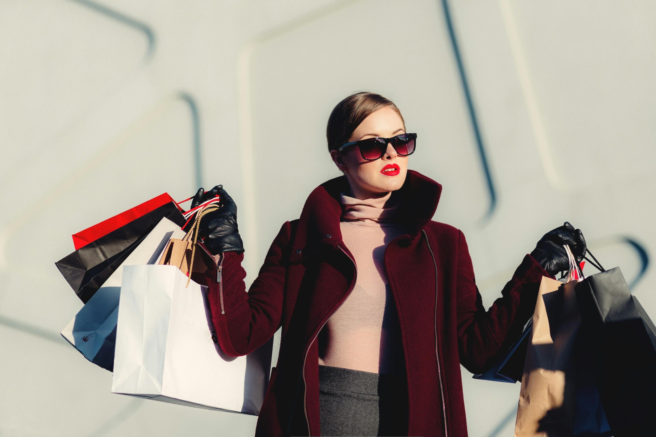 A woman in a burgundy coat wearing sunglasses is holding up a bunch of shopping bags.