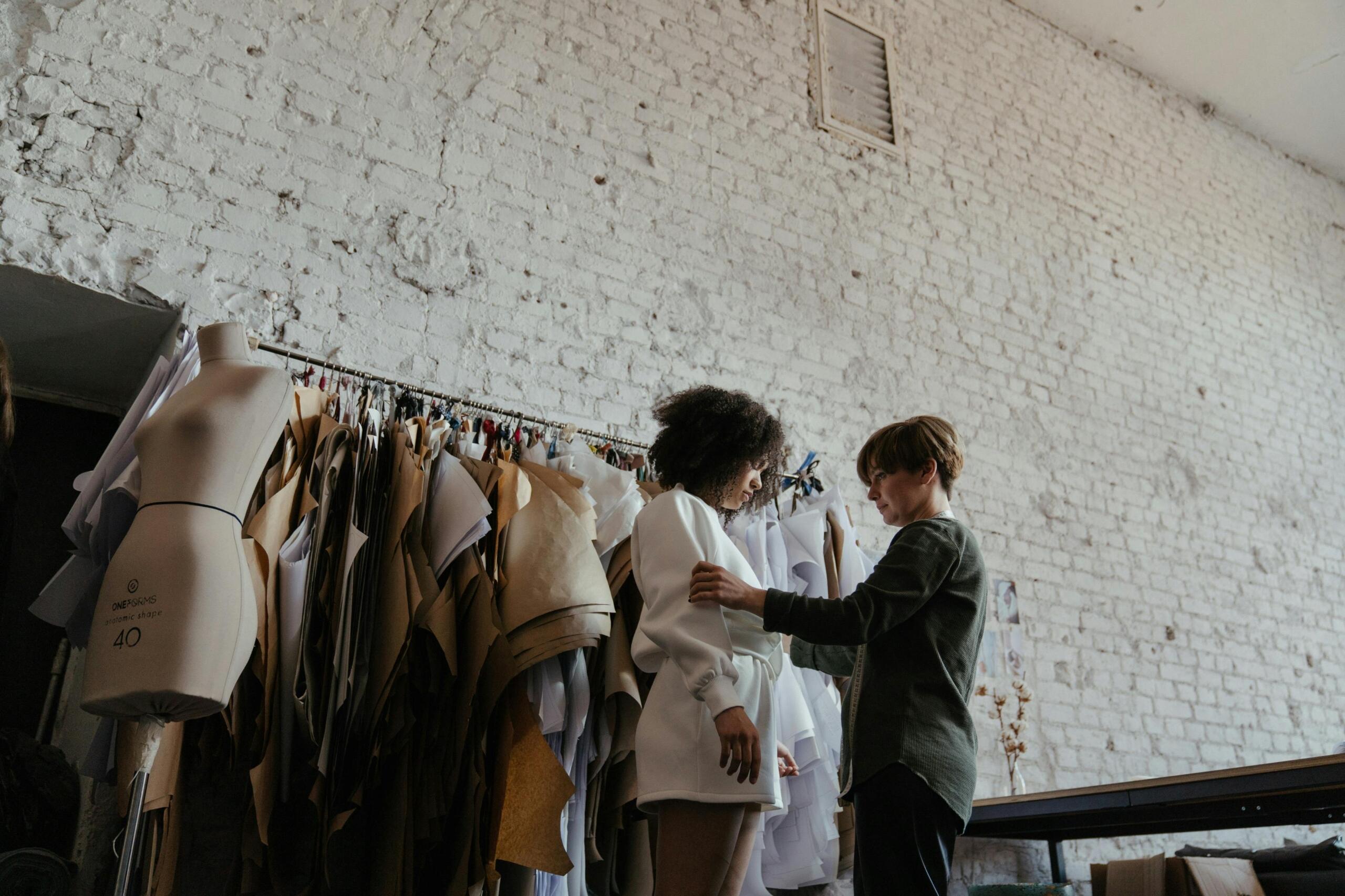 A fashion design student adjusts a garment on a model in a studio, surrounded by pattern papers and a dress form.