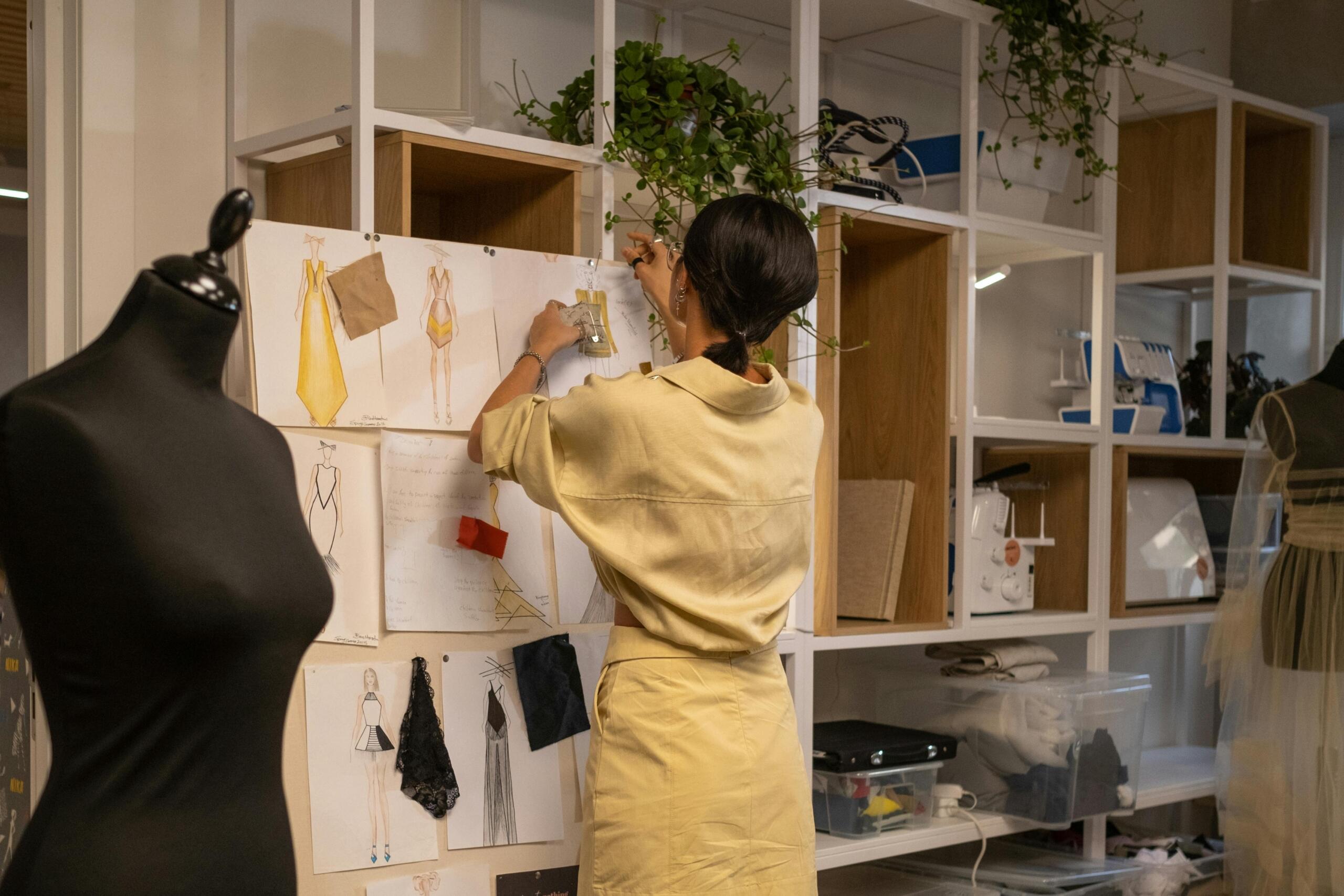 A woman in a beige outfit pins fabric swatches and fashion sketches to a mood board in a modern design studio, surrounded by mannequins, sewing machines, and shelving units with supplies.