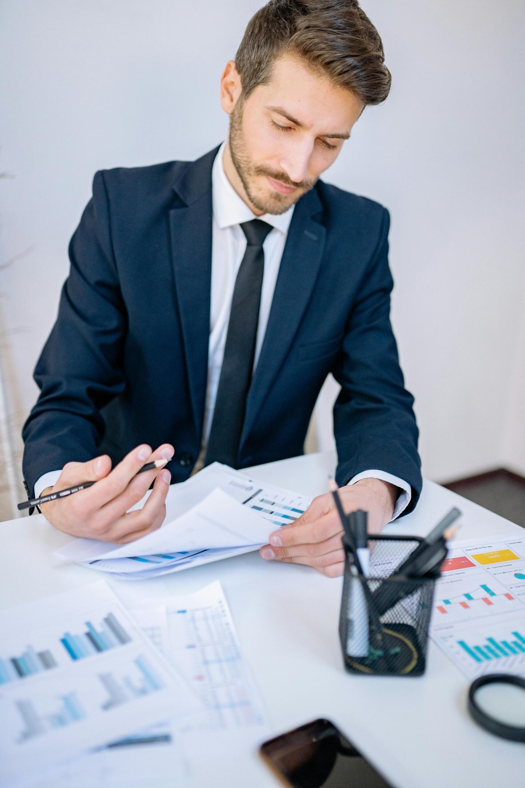 Financial analyst in a suit reviewing printed reports and charts at a desk, analyzing financial data and documents.