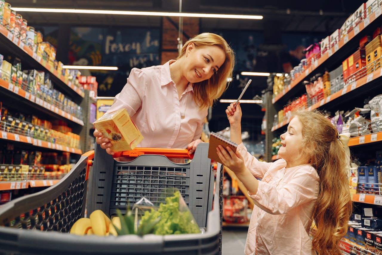 A woman and a girl shopping together in a grocery store, examining products in a shopping cart filled with fruits and vegetables.