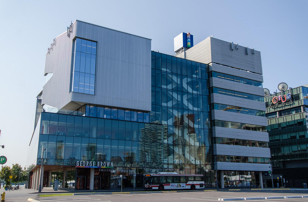 Modern glass and steel building of George Brown College in Toronto, with a city bus parked in front and high-rise reflections in the windows.