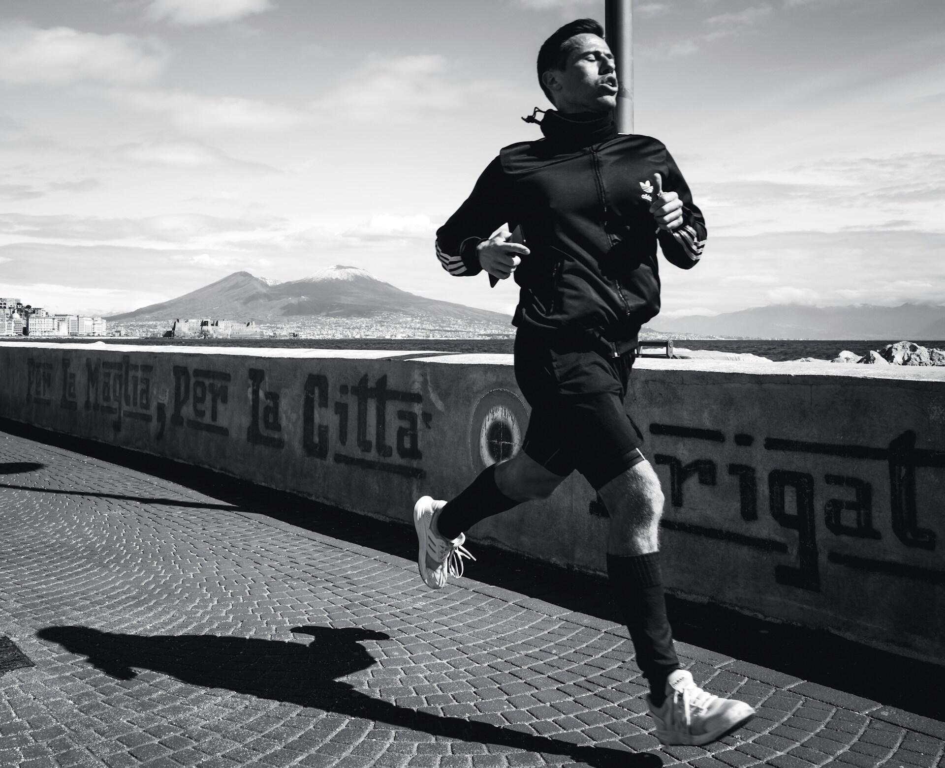 A runner in a black jacket and shorts jogs along a waterfront path with a view of a mountain under a cloudy sky.