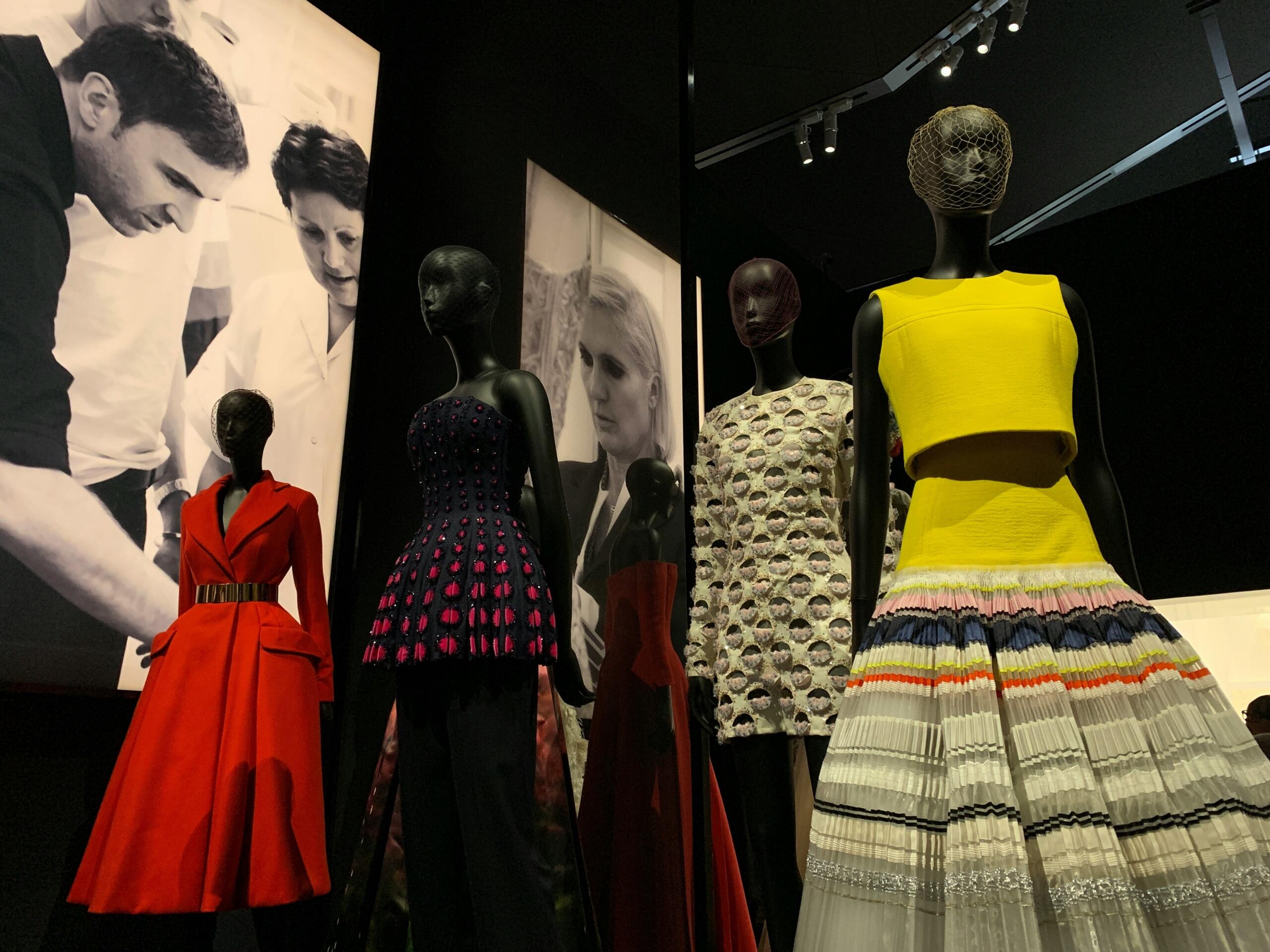 A display of five mannequins dressed in high-fashion garments, including a red coat dress, a yellow crop top with a pleated multicolored skirt, and intricately detailed dresses. The mannequins are set against a dark background with large black-and-white fashion photographs.