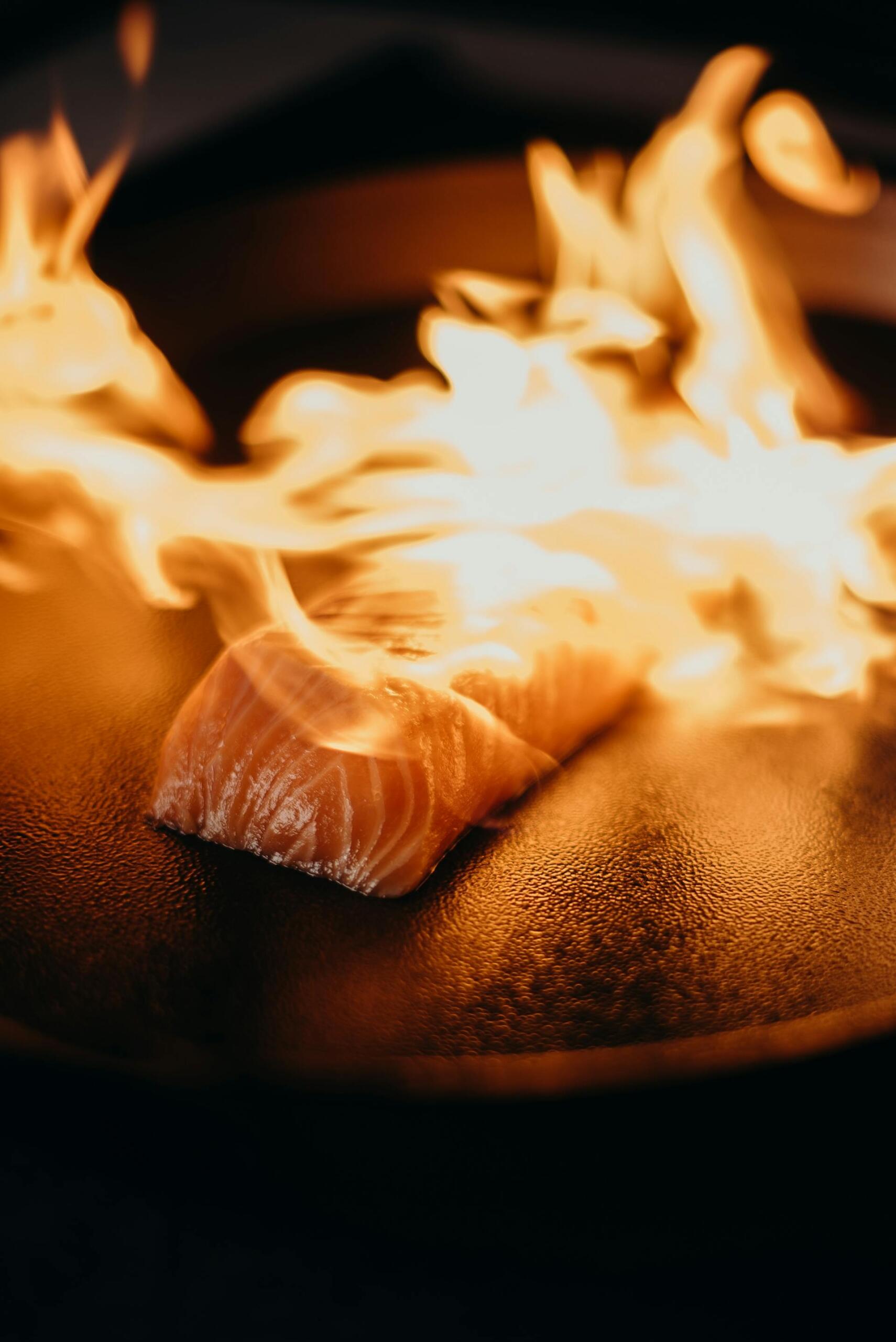 Flaming salmon fillet sizzling on a black grill, surrounded by vibrant orange and yellow flames, creating an intense cooking atmosphere.