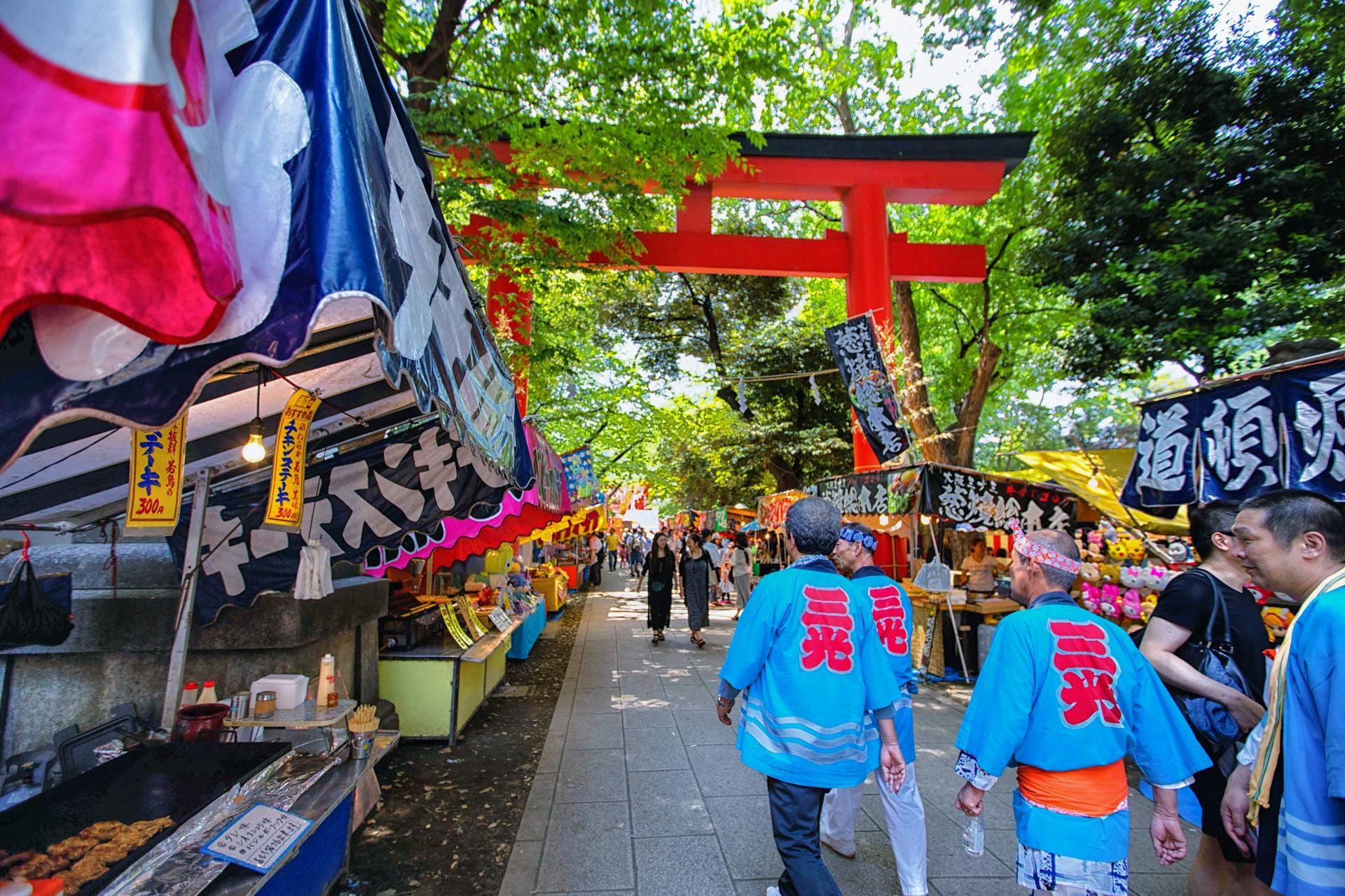 Colorful festival stalls line a shaded path, with banners and food vendors creating a vibrant atmosphere beneath green trees.