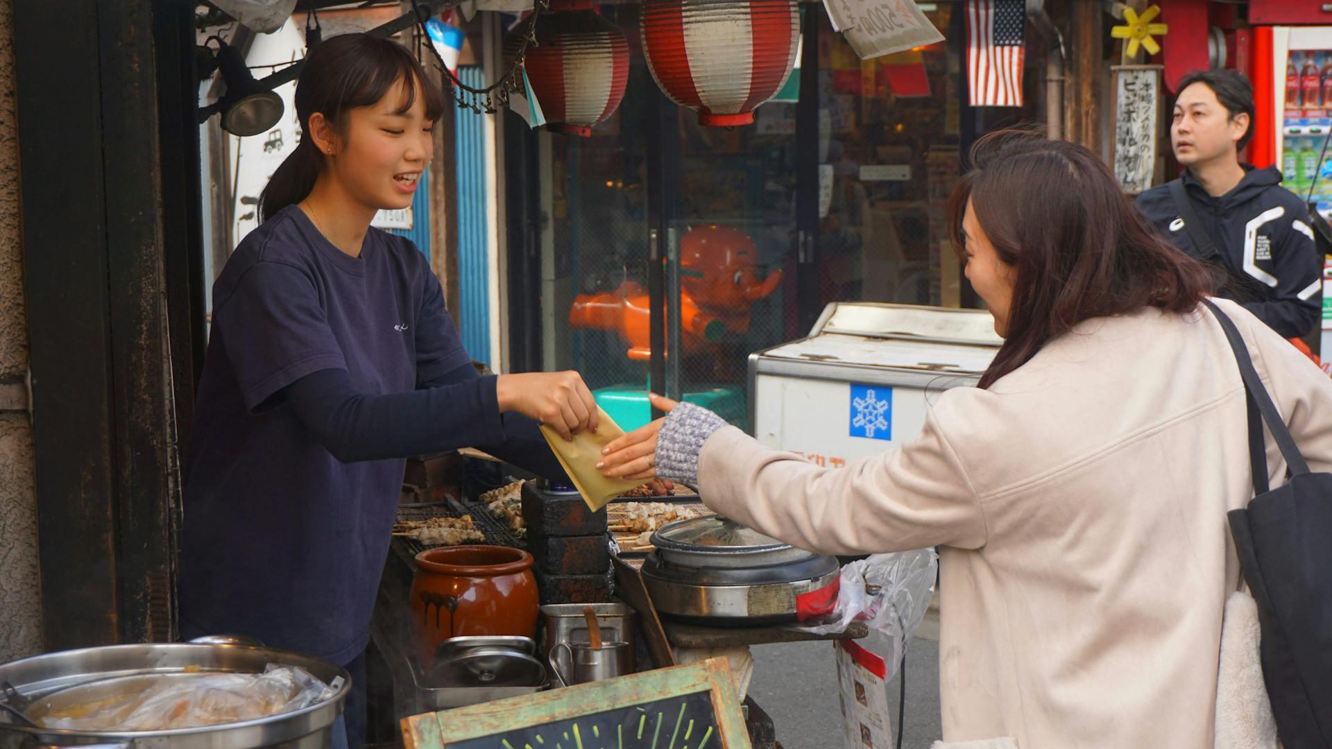 A vendor hands a wrapped food item to a customer at a street stall adorned with lanterns, surrounded by colorful shops.