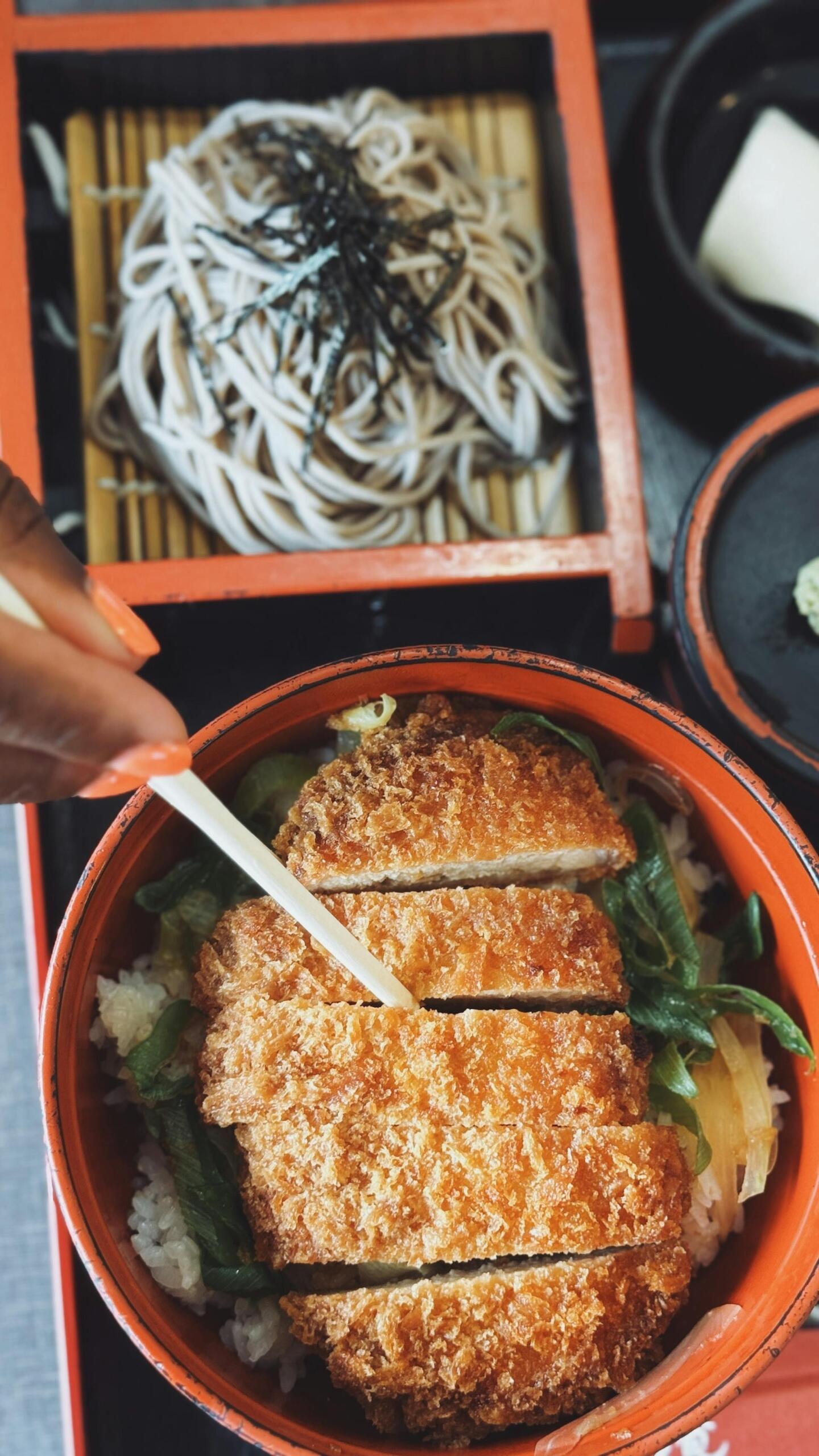 A hand using chopsticks to cut a crispy pork cutlet in a bowl, alongside a side of soba noodles and dipping sauce.