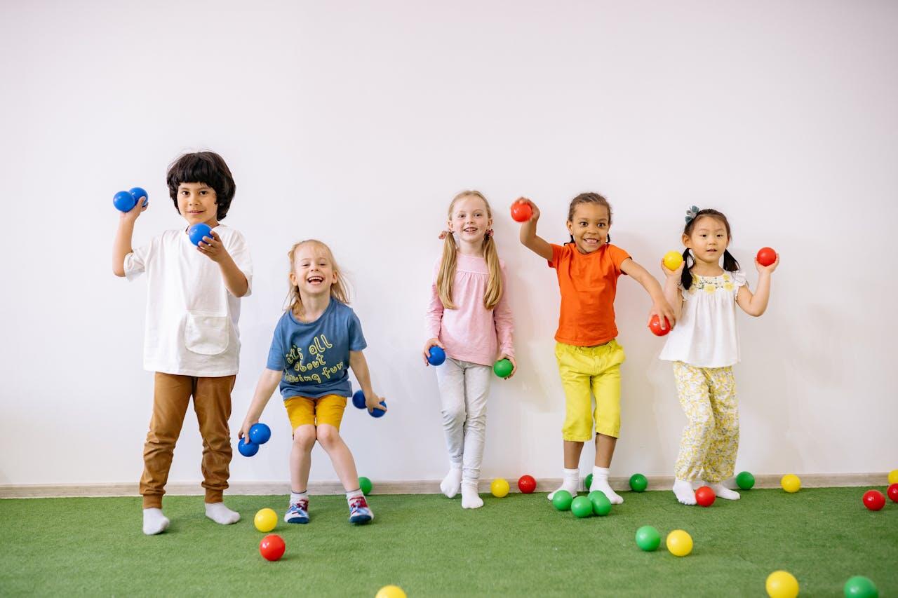 Five children stand in a row, holding colorful dumbbells, surrounded by scattered playful balls on a green carpeted surface.