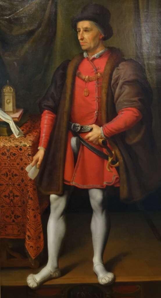 A man in a red tunic and brown fur cloak stands confidently, holding a small object, beside a table adorned with a patterned cloth.