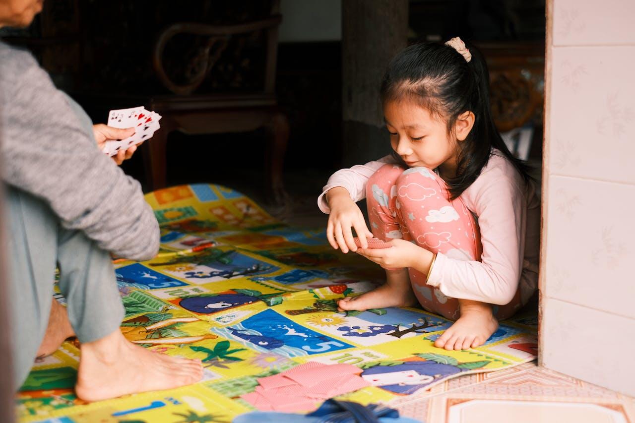A child with long hair sits on a colorful mat, focused on a card game being played by an adult nearby.