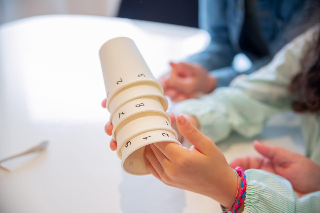 A child’s hand holds a stack of numbered white cups, while another hand reaches toward them, suggesting a play or learning activity.