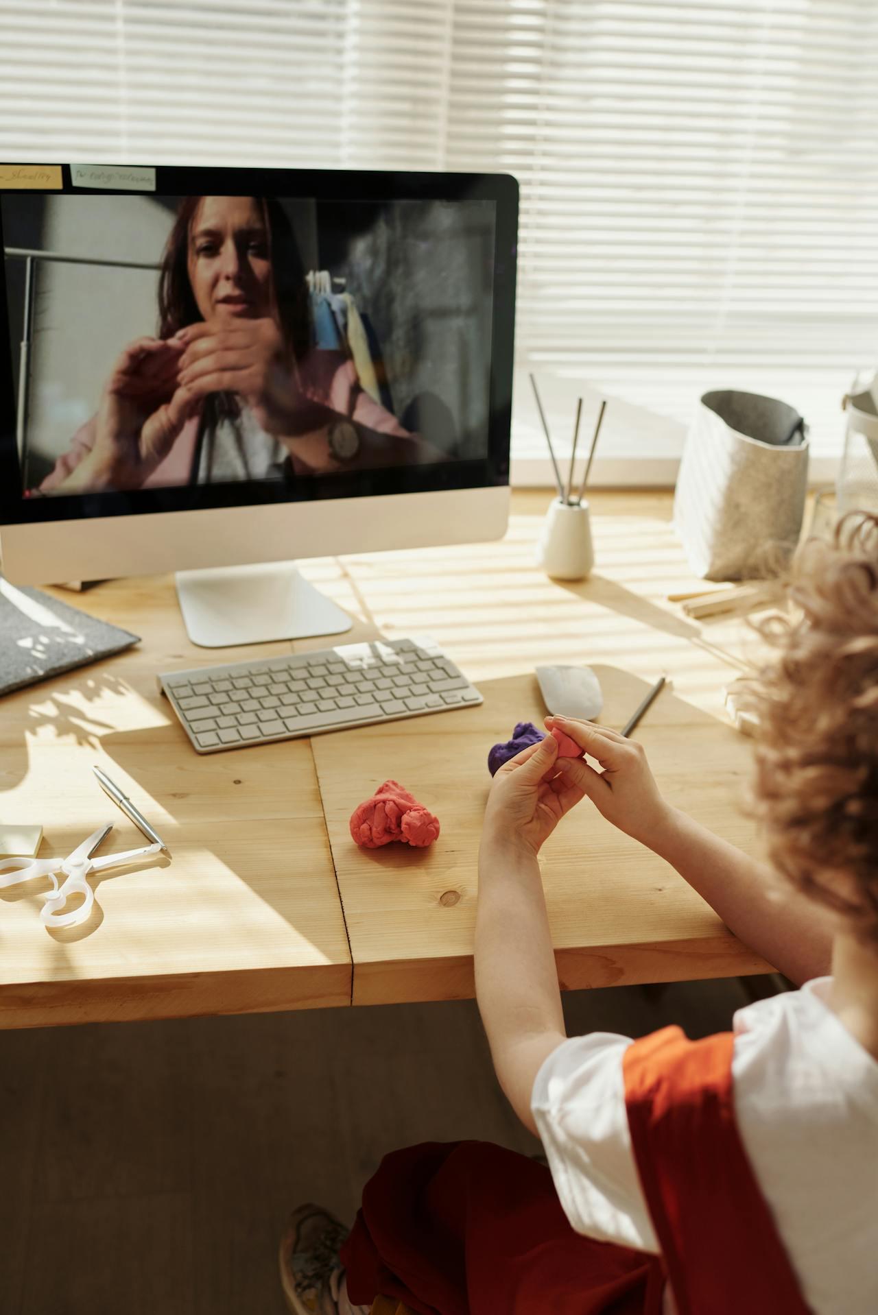 A child sits at a wooden desk, crafting with colorful clay while engaging with a video call on a computer screen.