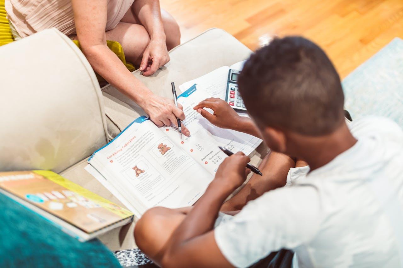 A child and an adult work together on a homework task, pointing at a workbook filled with illustrations and exercises on a couch.