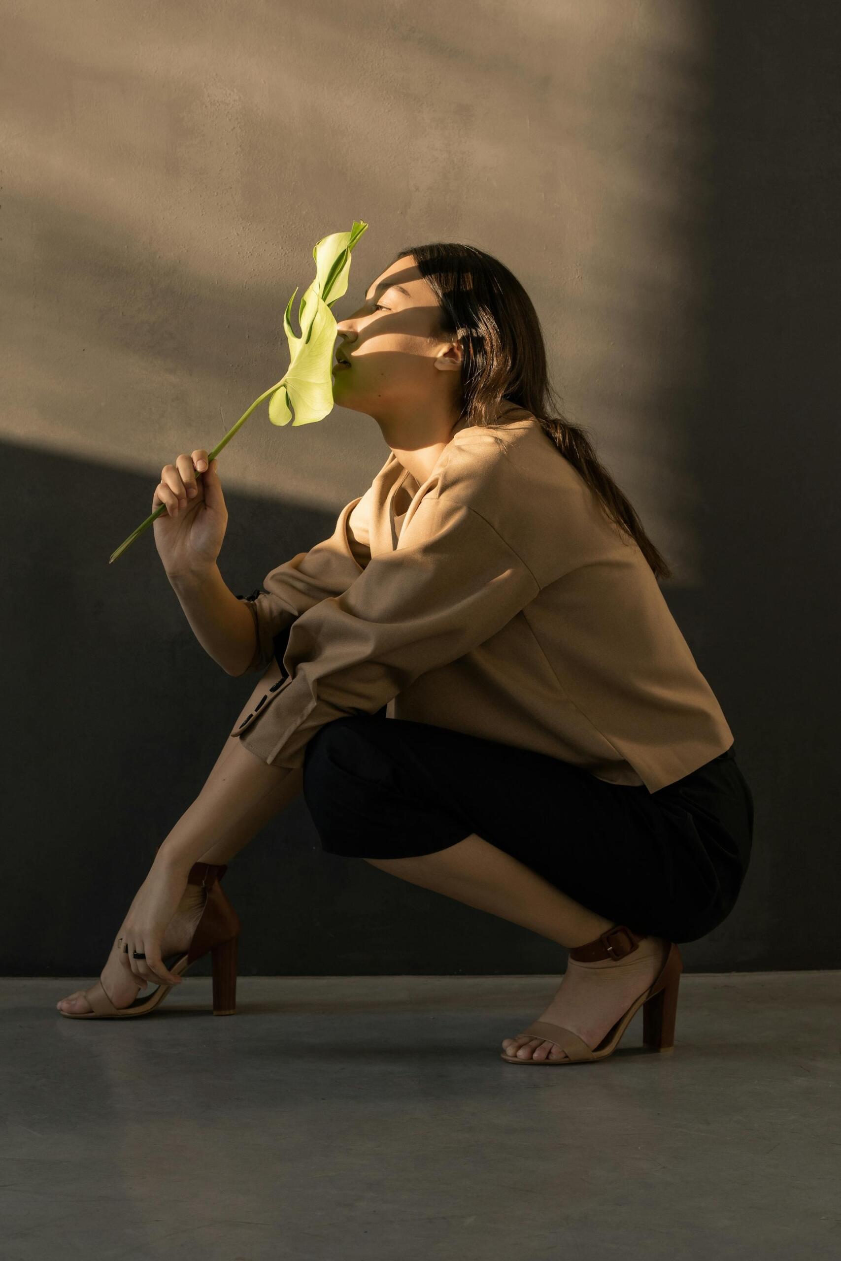 A person squats against a dark wall, holding a large green leaf, dressed in a beige top and black pants with stylish high heels.