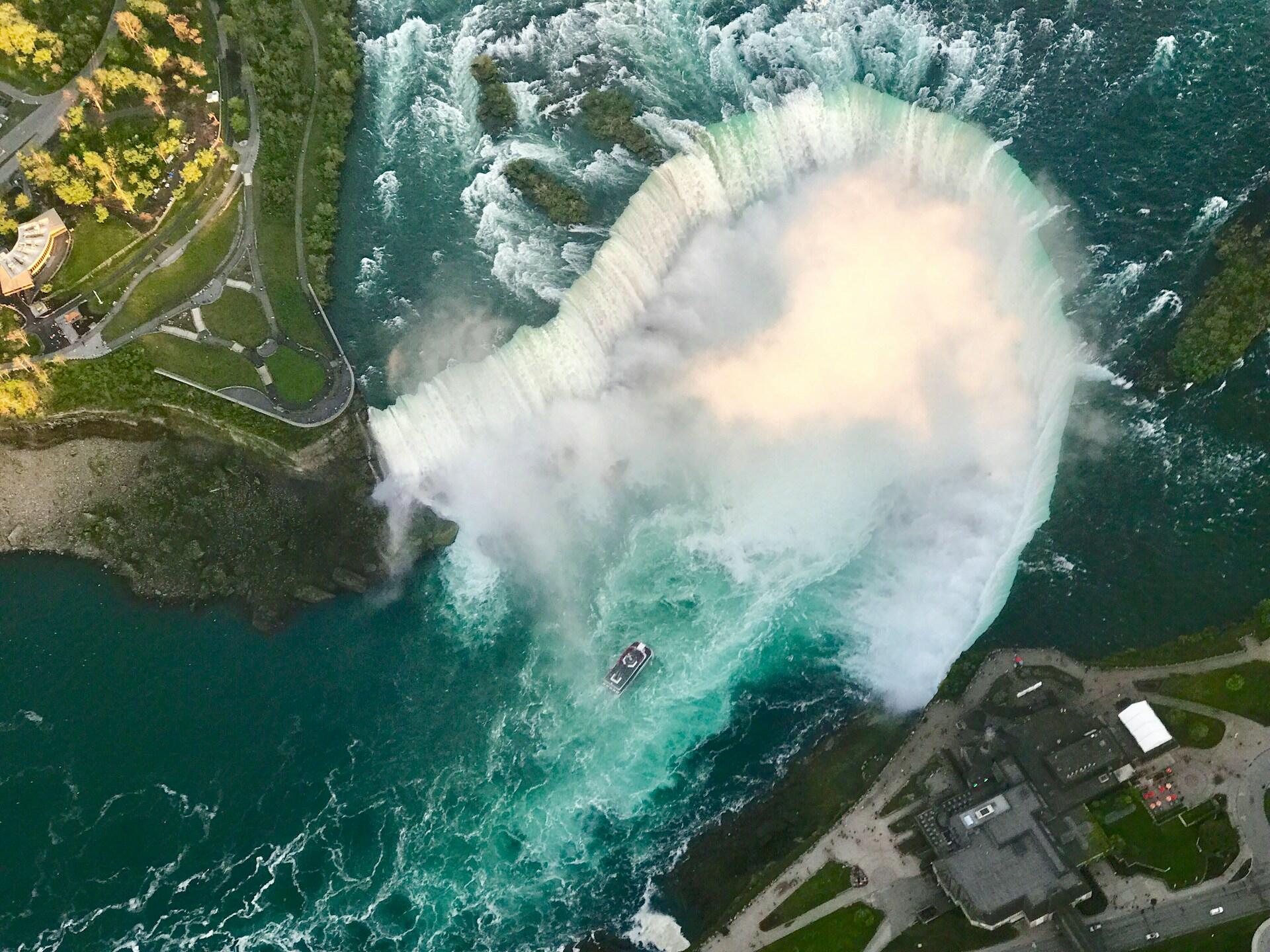 Aerial view of Niagara Falls, showcasing cascading water, mist, a boat near the base, and lush green surroundings.