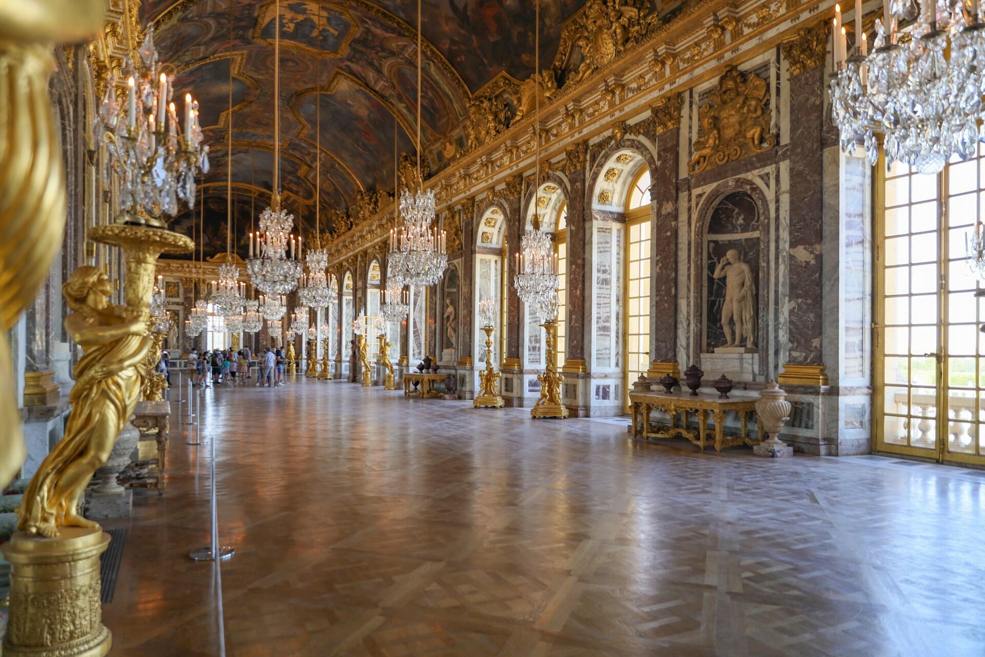 Interior of the Hall of Mirrors at Versailles, showcasing opulent chandeliers, golden sculptures, and ornate decor under a painted ceiling.