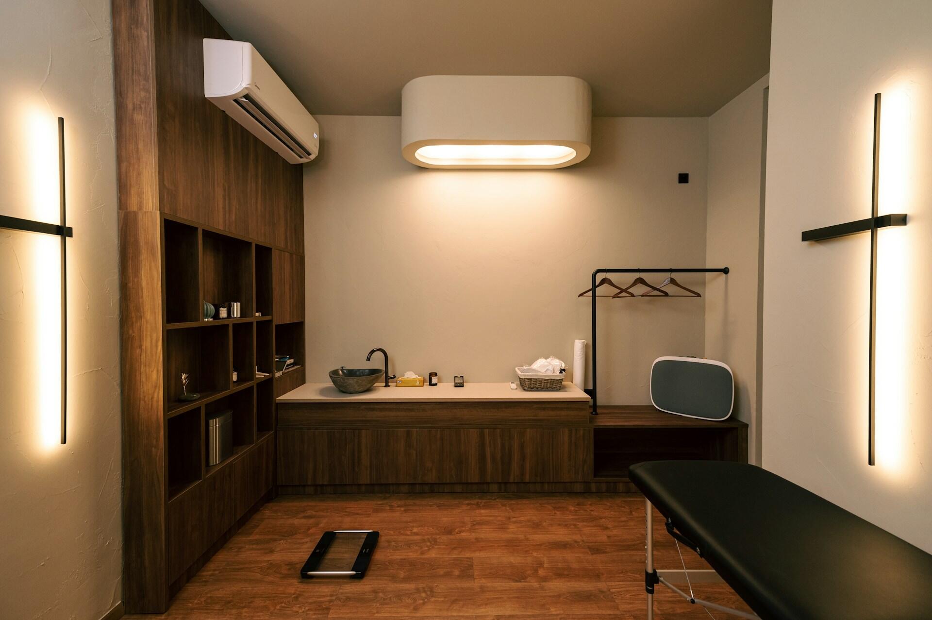 A minimalist treatment room featuring wooden shelves, a sink, a treatment table, and soothing lighting for a calming atmosphere.