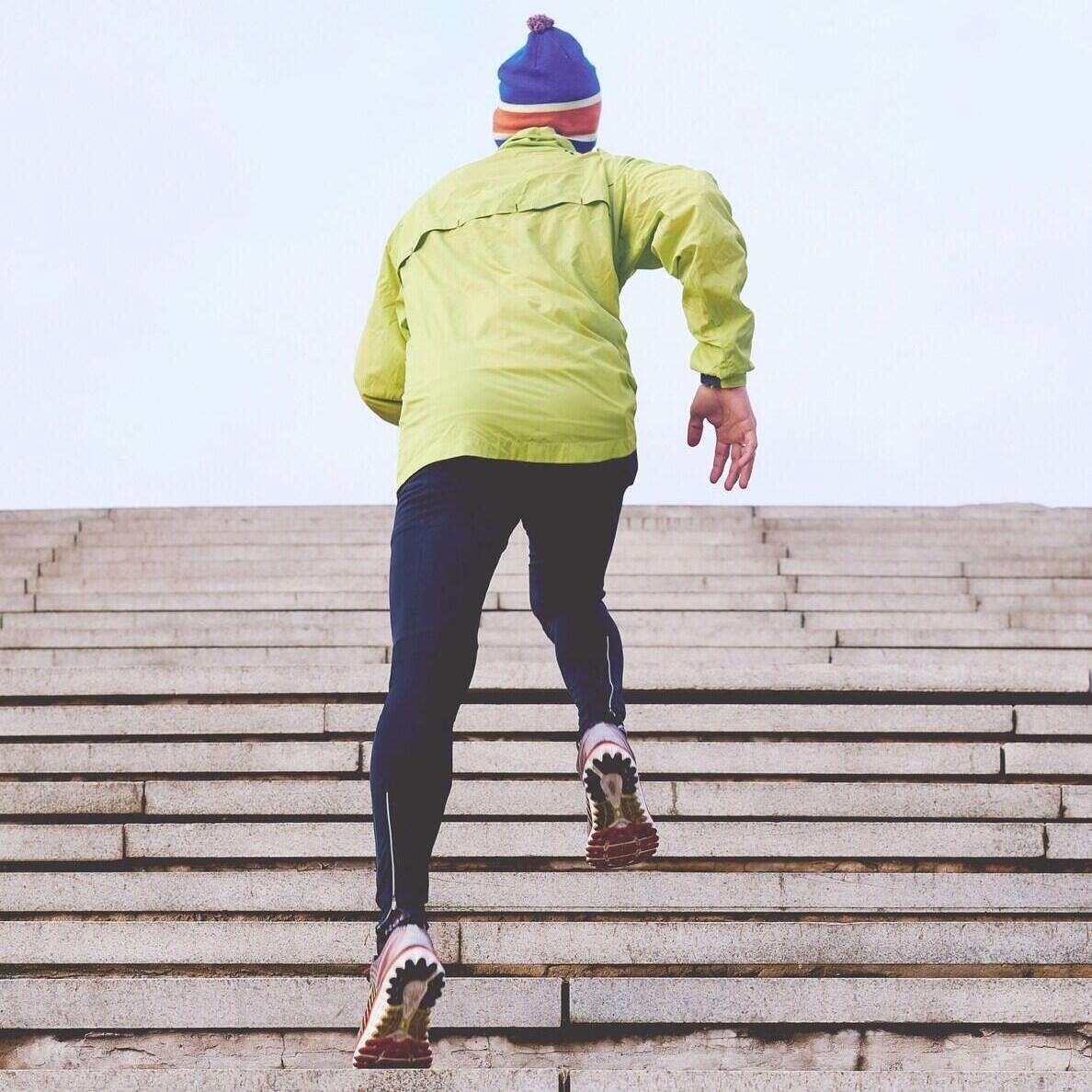 A person in a bright yellow jacket and blue hat runs up a flight of outdoor stairs, viewed from behind.