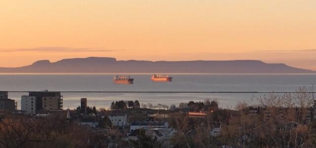Two cargo ships anchored on calm waters at sunset, with a distant mountain silhouette and a town visible in the foreground.