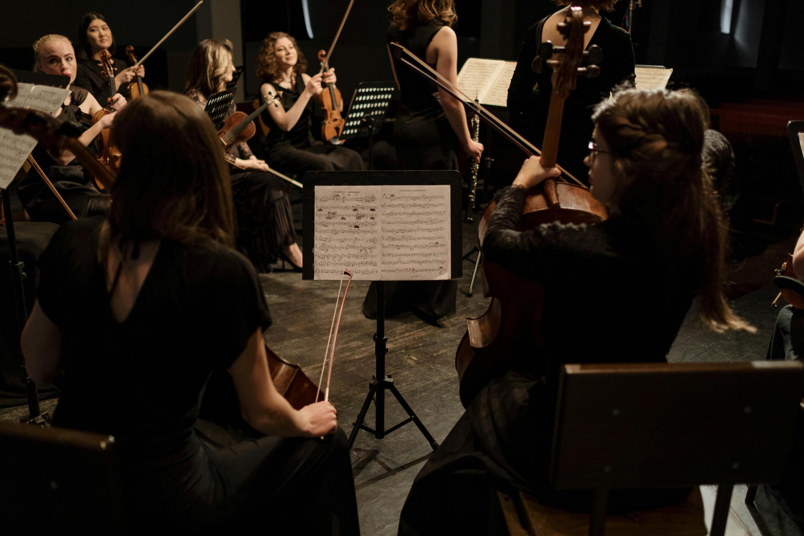 A group of string musicians sit in a circle with their sheet music in front of them.