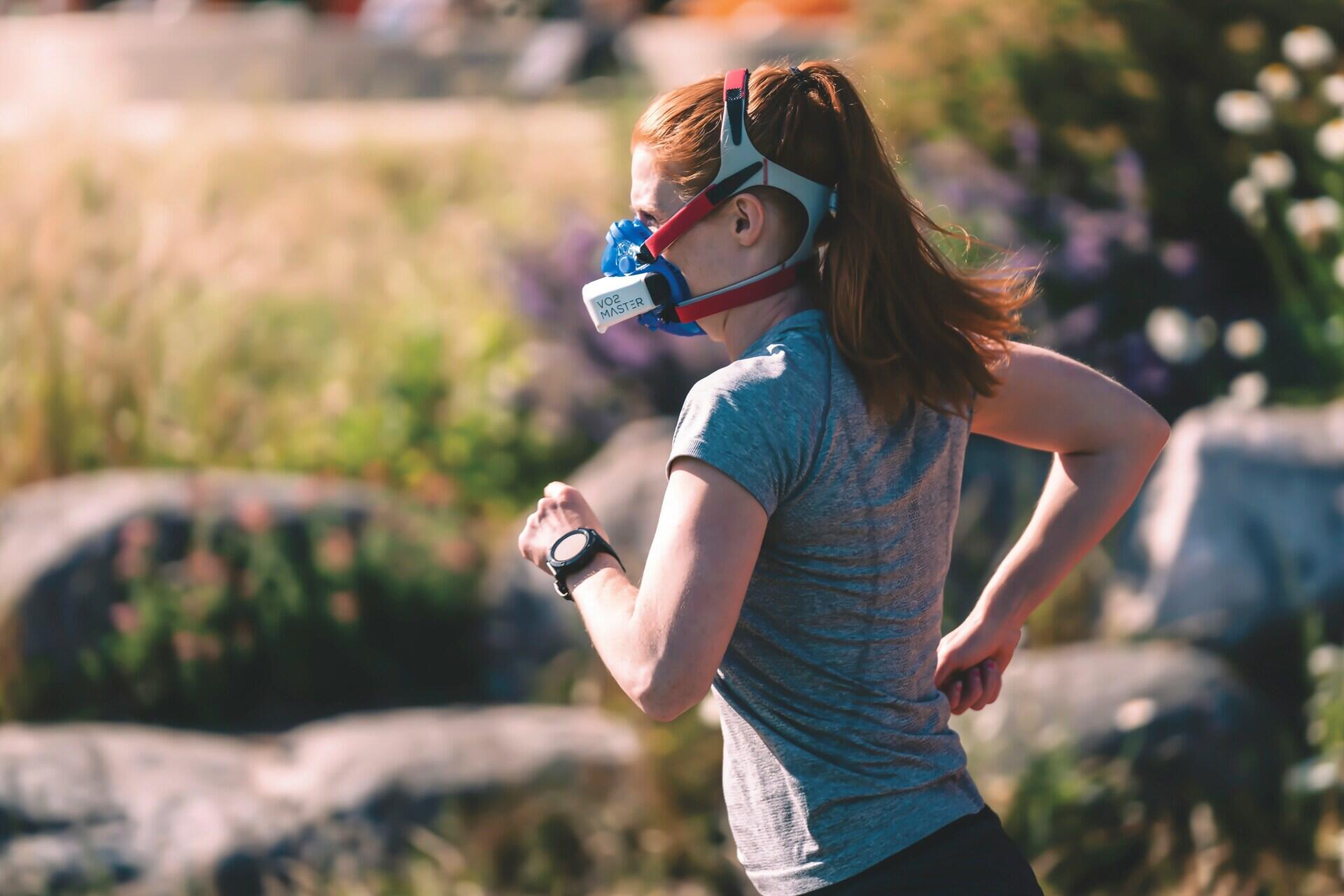 A woman in a gray t-shirt runs outdoors, wearing a fitness tracker and a face mask, surrounded by greenery and vibrant flowers.