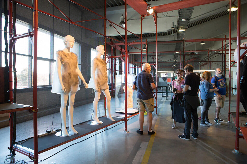 Two mannequins dressed in translucent, textured garments are displayed on a raised platform inside an industrial-style room with red metal scaffolding. Several people stand nearby observing and discussing the exhibit. Large windows line the left side of the space, letting in natural light.
