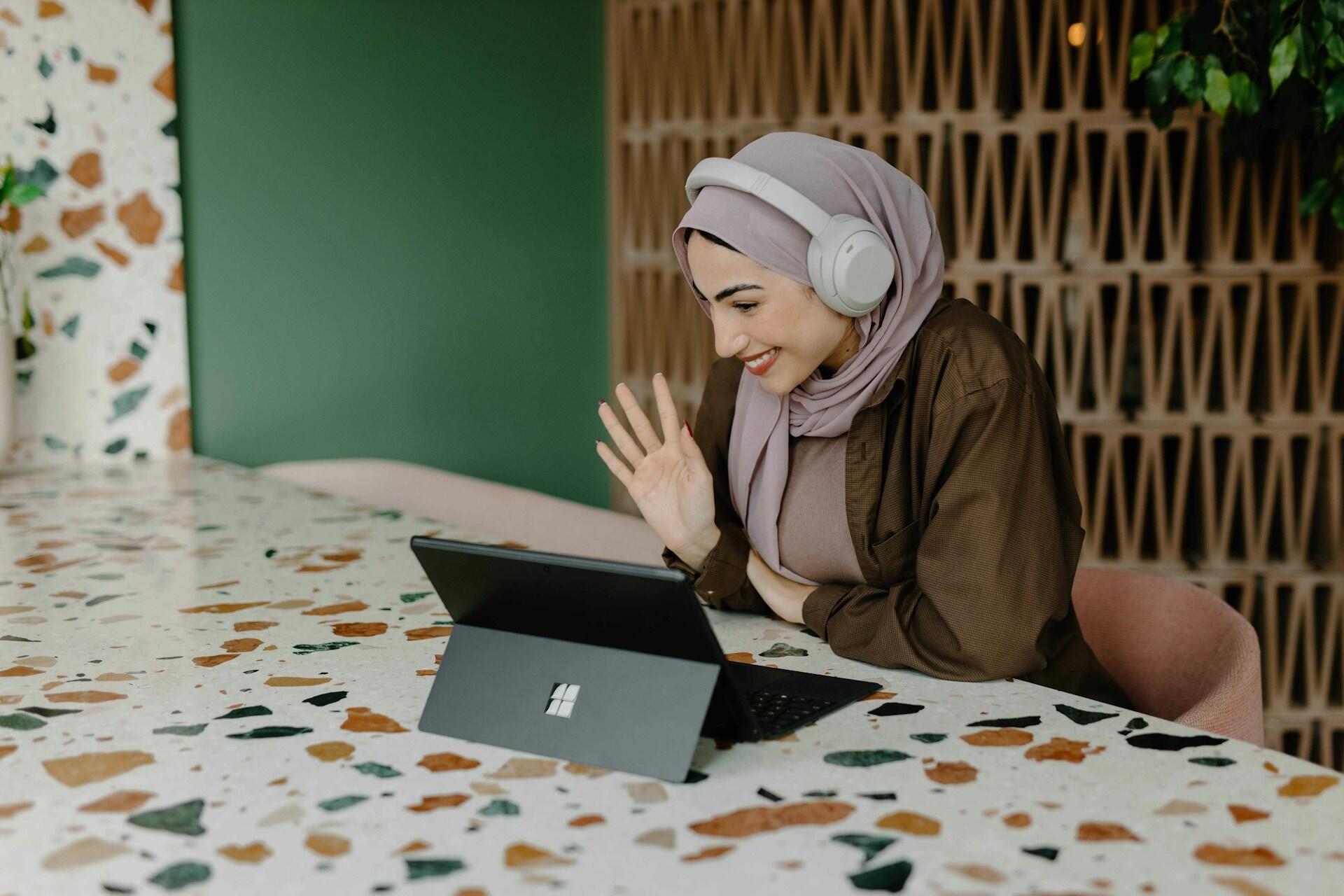 A woman in a hijab wearing headphones is sitting at a terrazzo table, engaging with a tablet in front of her.
