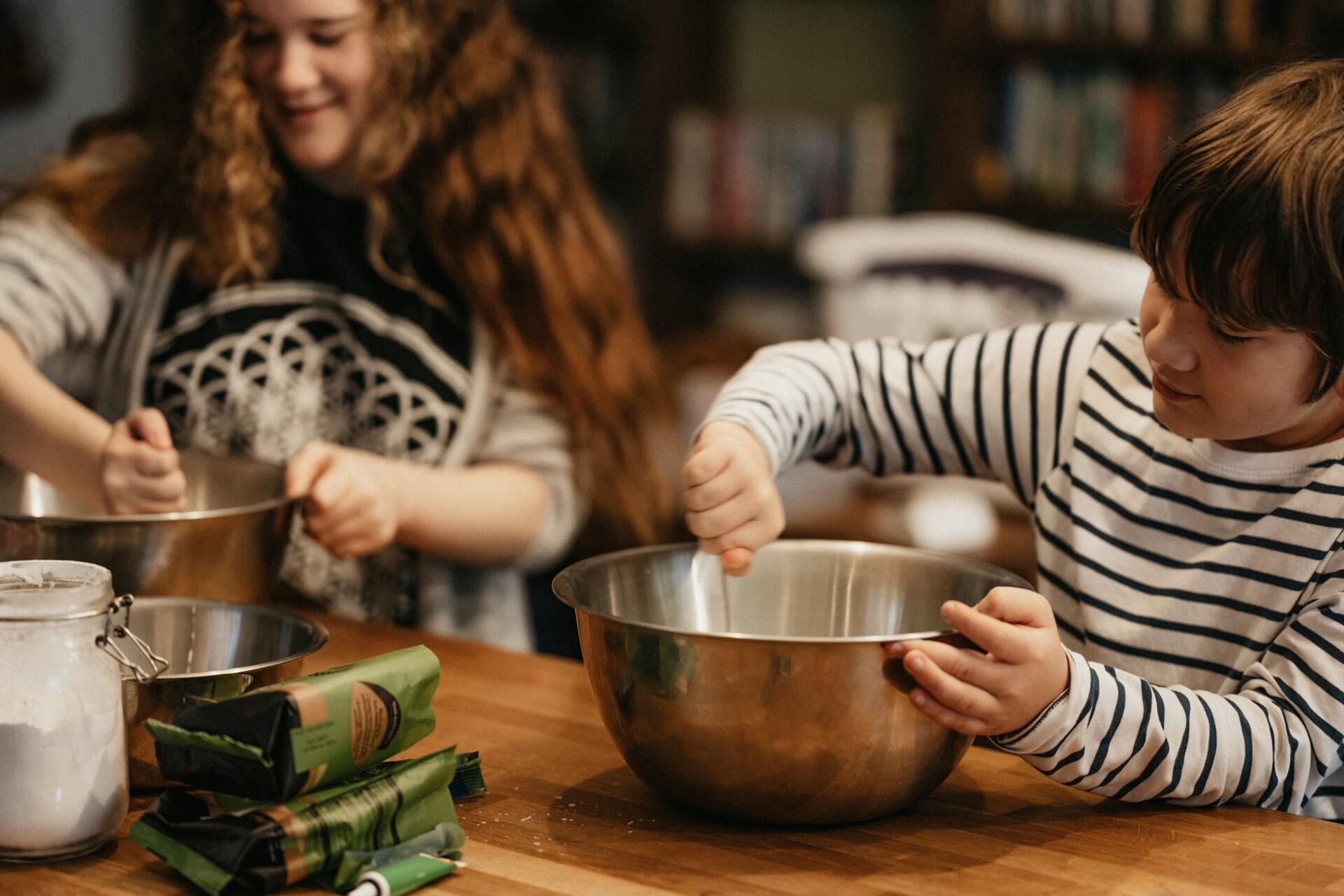 Children mixing ingredients in stainless steel bowls, surrounded by baking supplies on a wooden counter in a cozy kitchen.