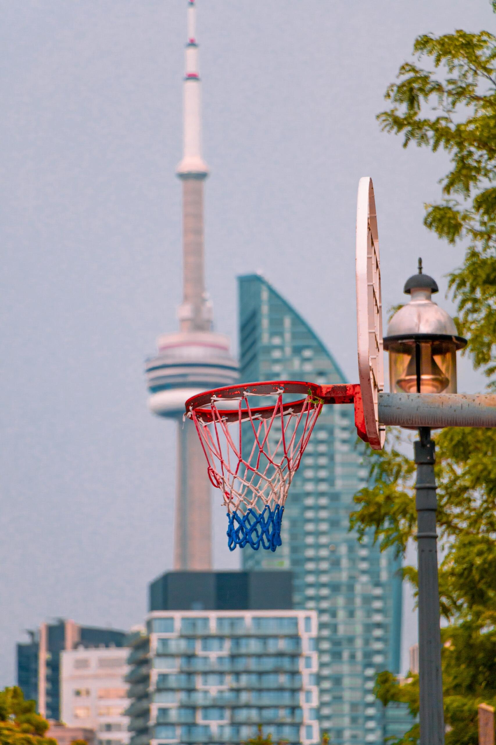 A close-up of a basketball hoop with a vibrant net, set against the Toronto skyline featuring the CN Tower and modern buildings.