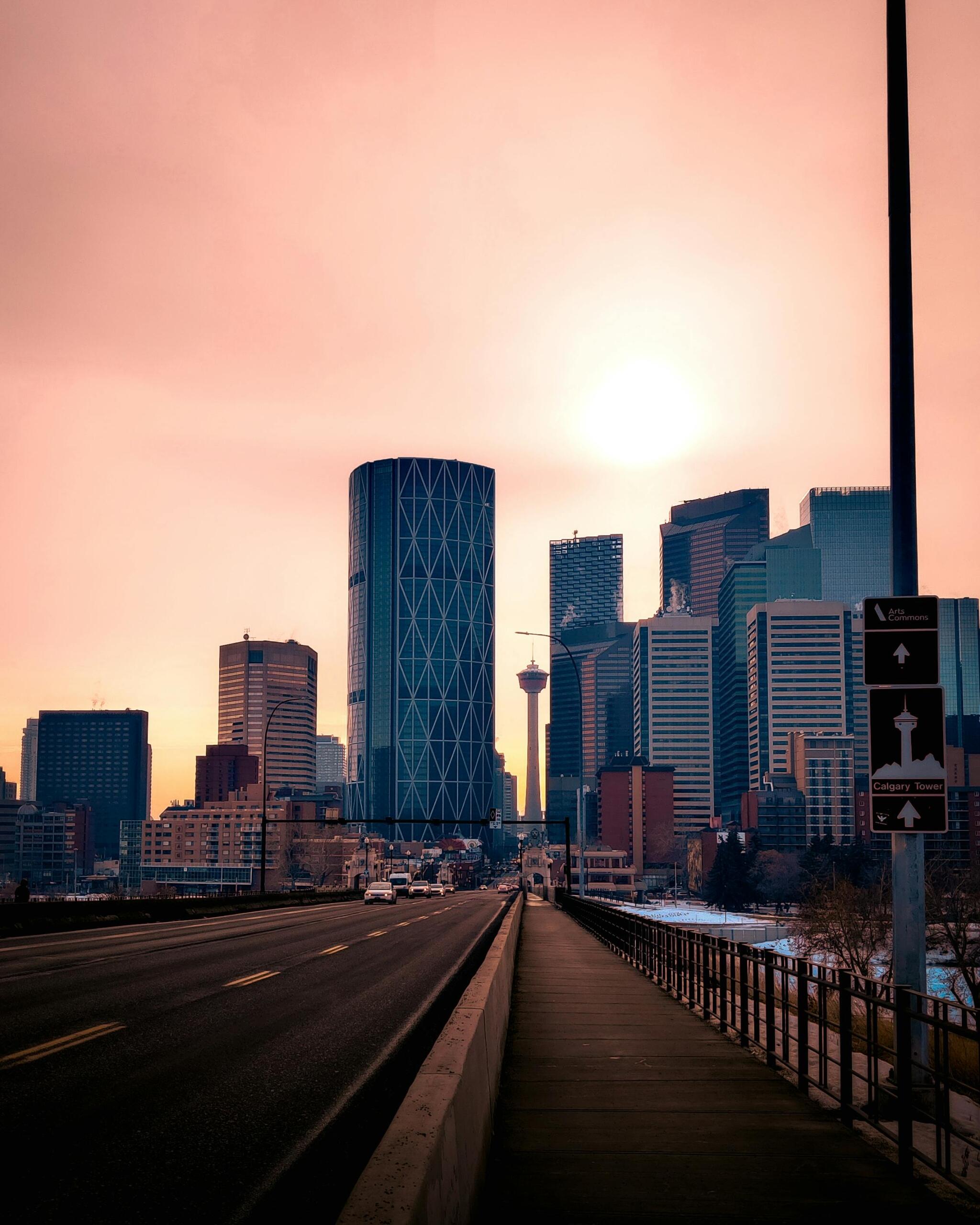 A view of downtown Calgary, showcasing modern skyscrapers, streets, and the surrounding cityscape under a pink sky. Source: Dilusha Gonagala.