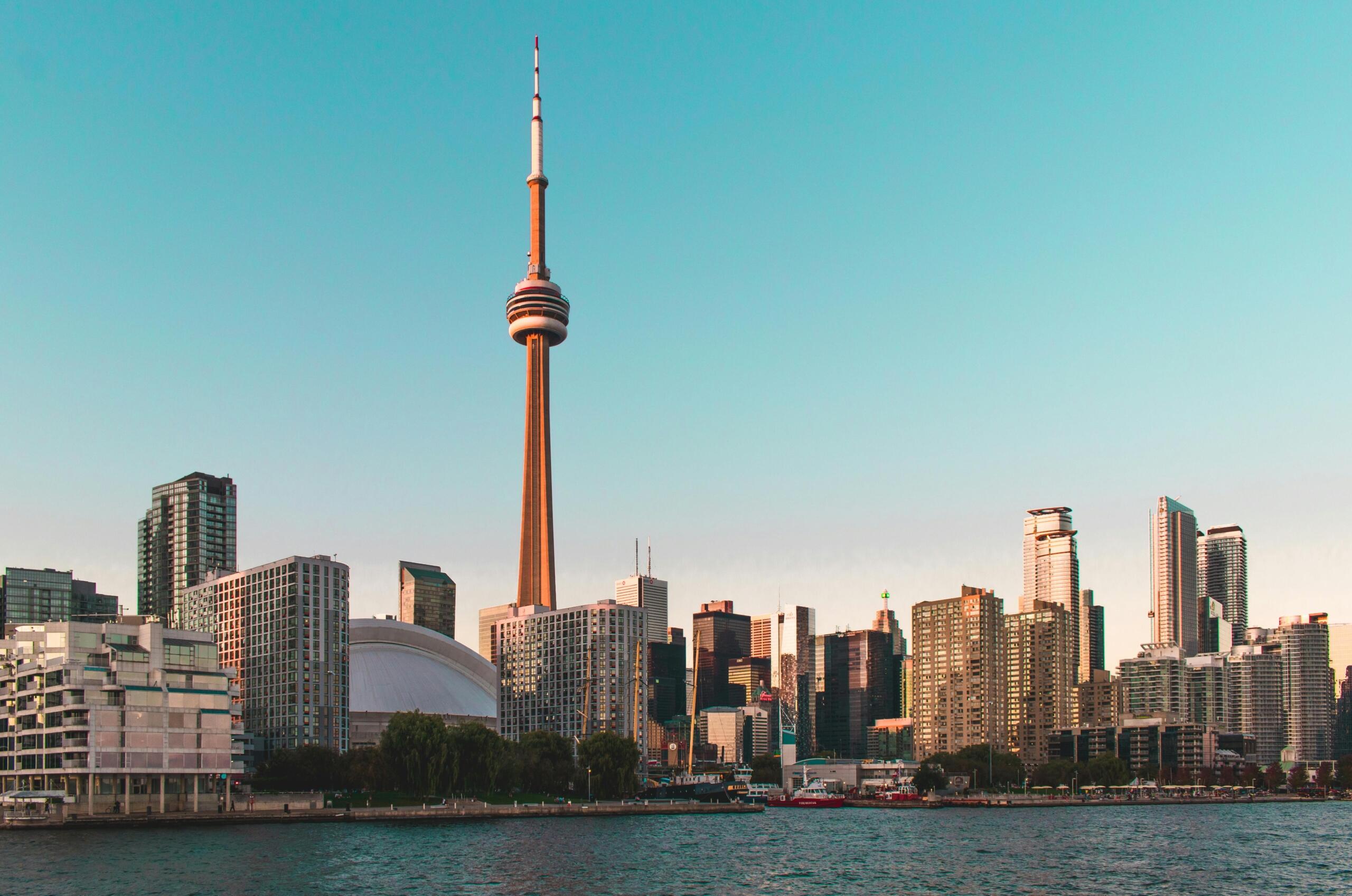 The Toronto skyline featuring the CN Tower surrounded by modern high-rise buildings, with Lake Ontario in the foreground and a clear sky overhead.