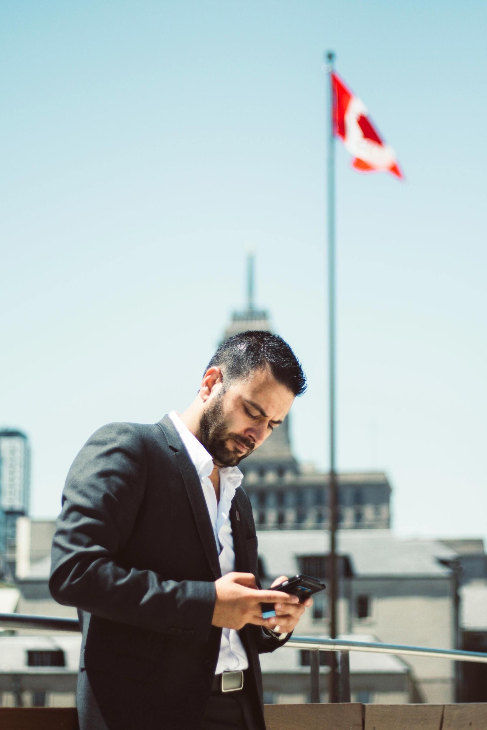 A man in a suit standing in front of a Canadian flag, checking his phone with a focused expression. Source: Yama Salghani.