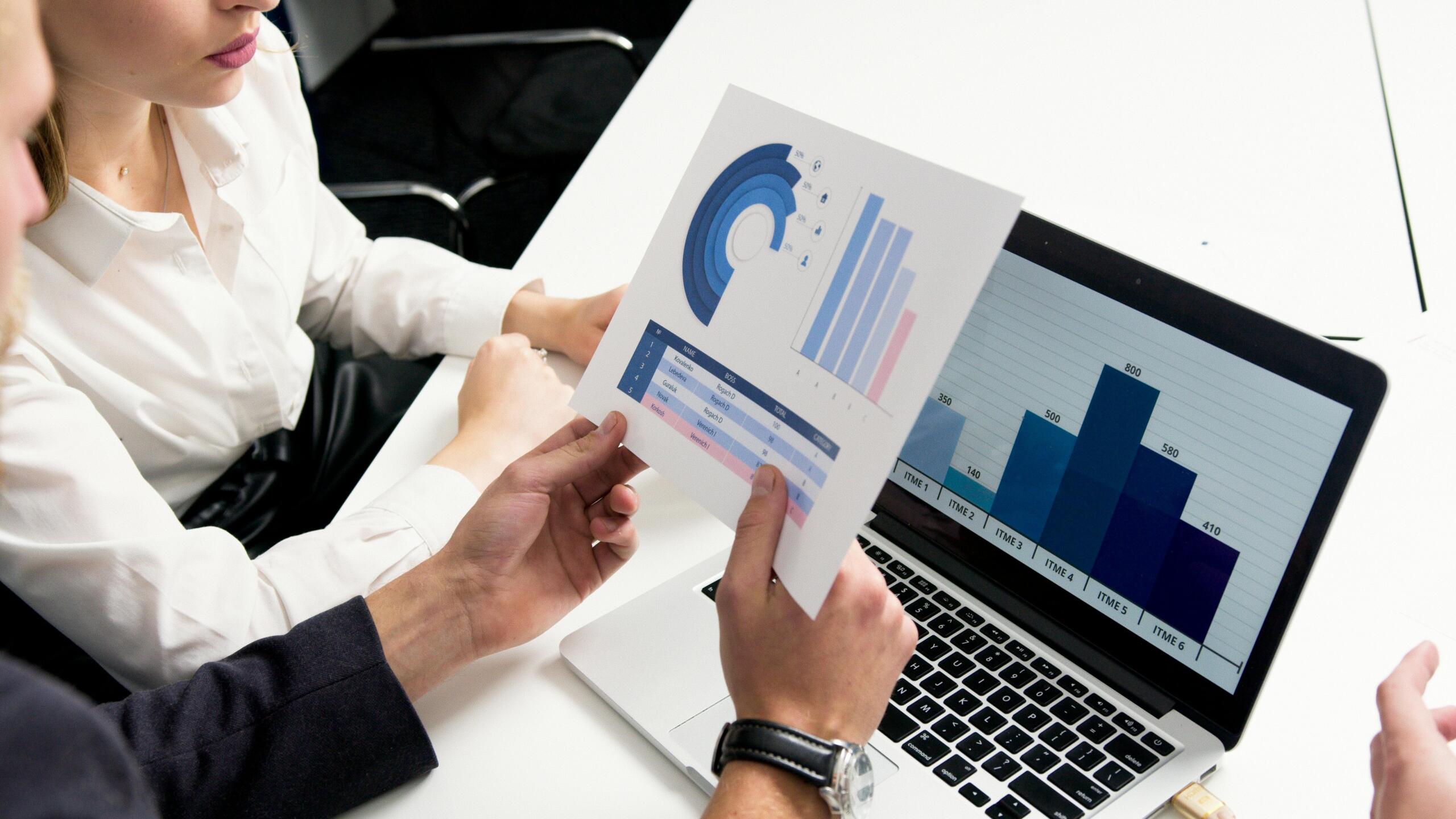 Close-up of two professionals’ hands by a laptop, holding printed charts and discussing data during a meeting.