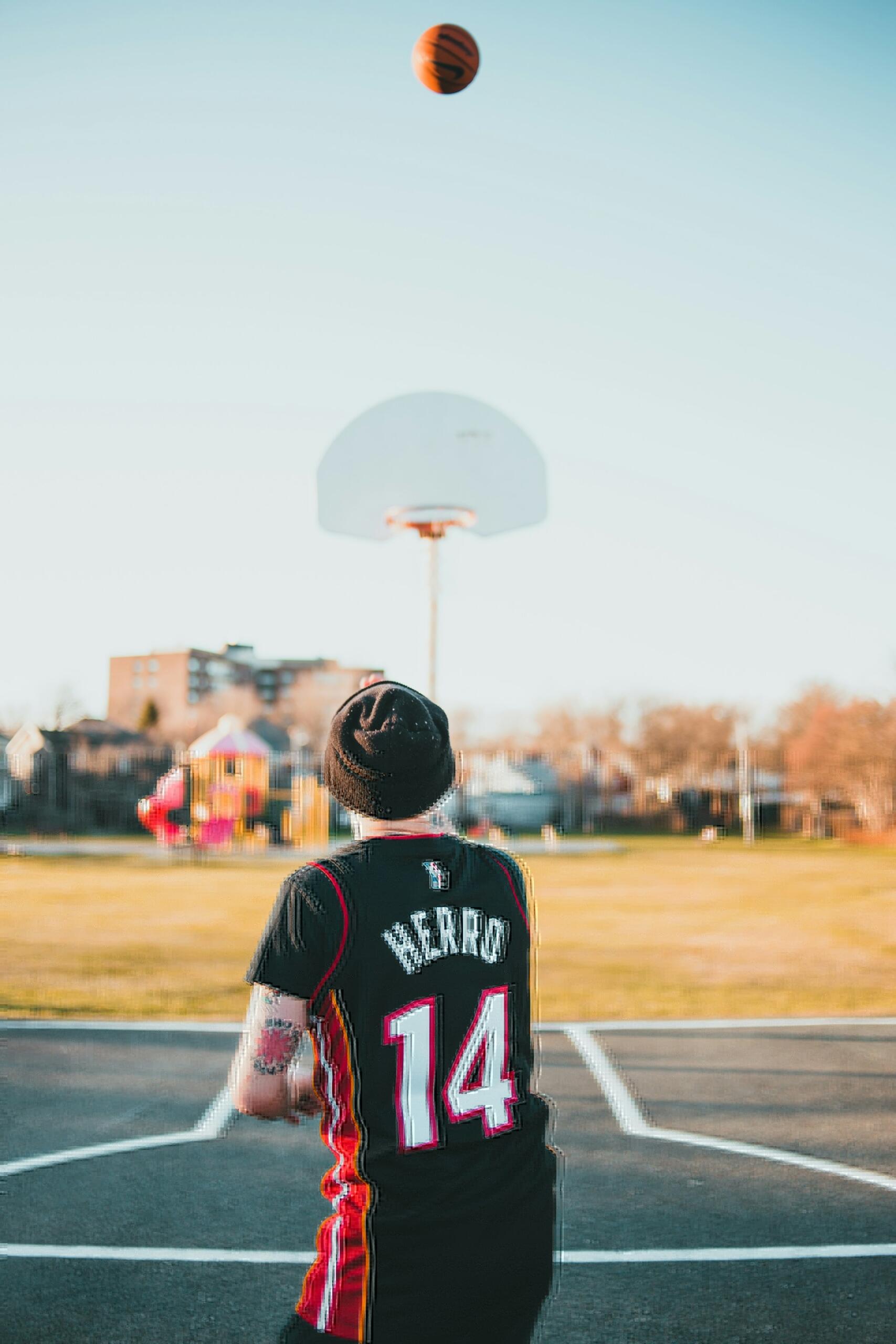 A person in a black basketball jersey with "HERRO" on the back shoots a basketball towards a hoop in a sunny park.