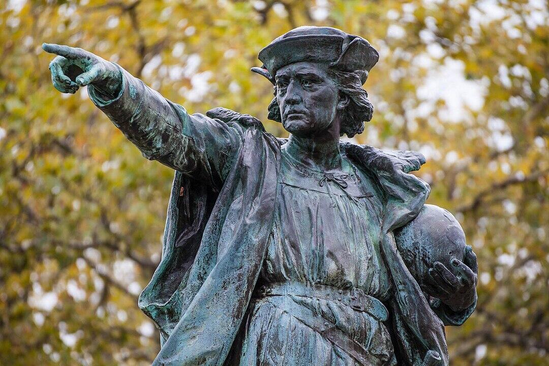 A weathered bronze statue of a historical figure, raised hand pointing, holding an orb in the other hand, surrounded by autumn foliage.