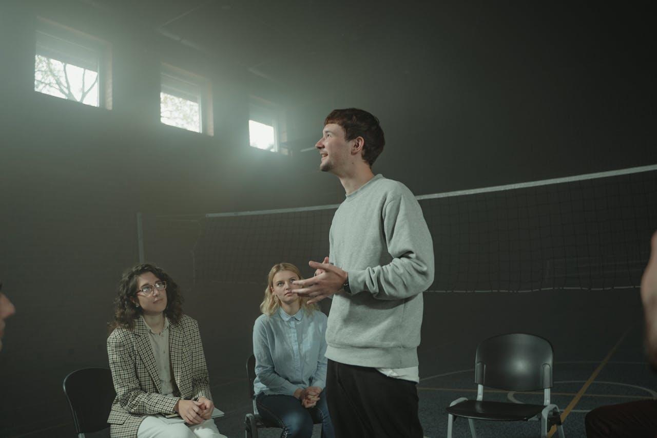 A person stands and speaks in a dimly lit gym, while three individuals seated attentively listen. A volleyball net is visible in the background.