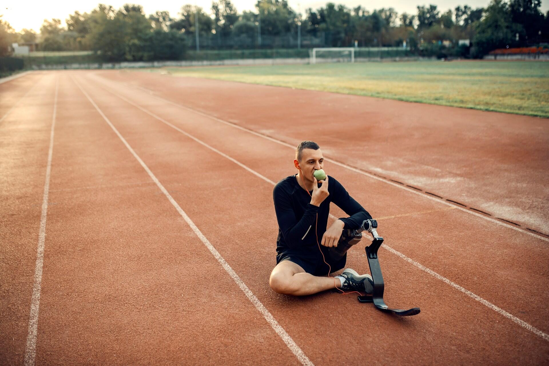 An athlete sits on a running track, using a prosthetic leg, appearing contemplative in a serene outdoor setting.