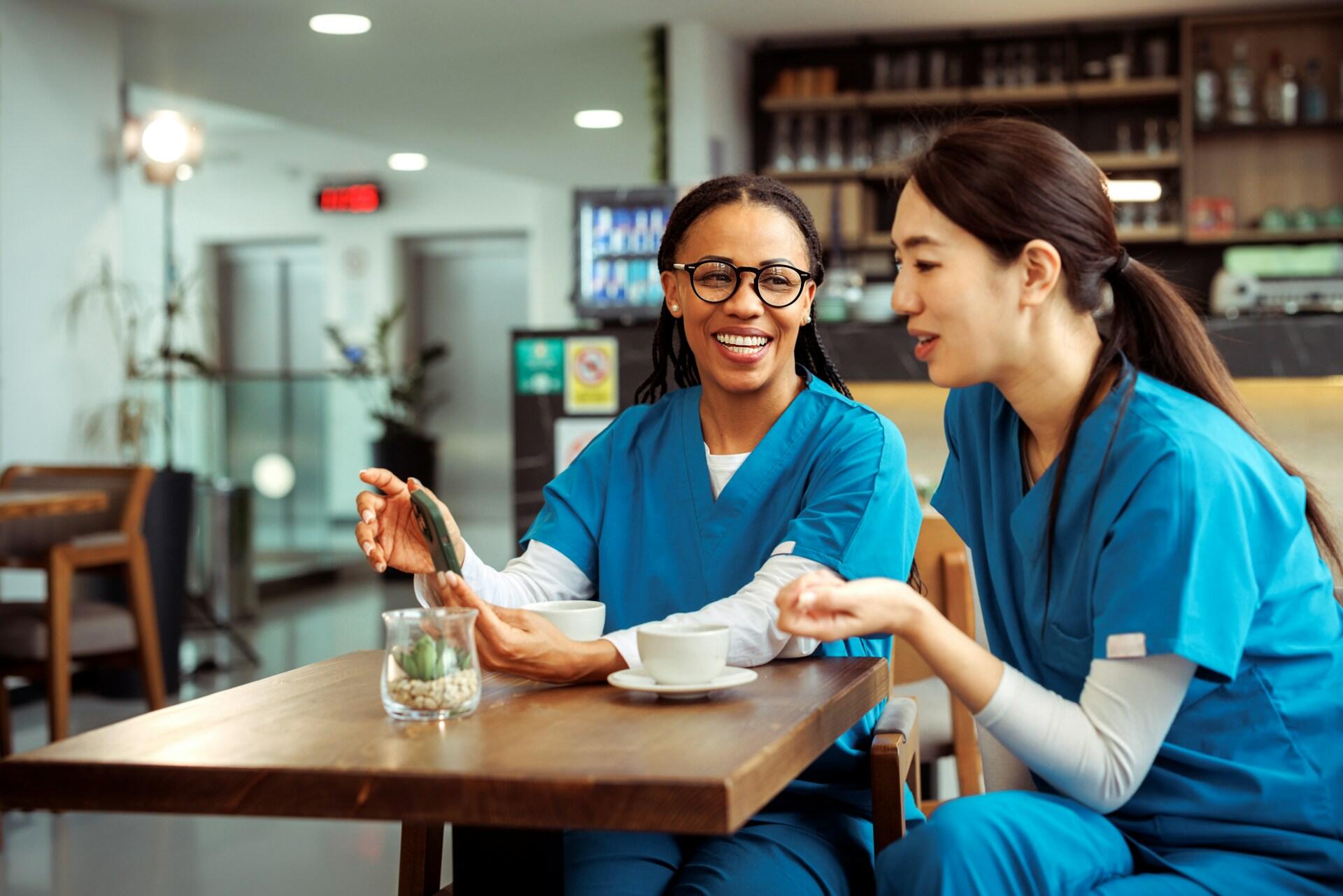 Two healthcare professionals in scrubs share a conversation over coffee at a café, with a small plant on the table.