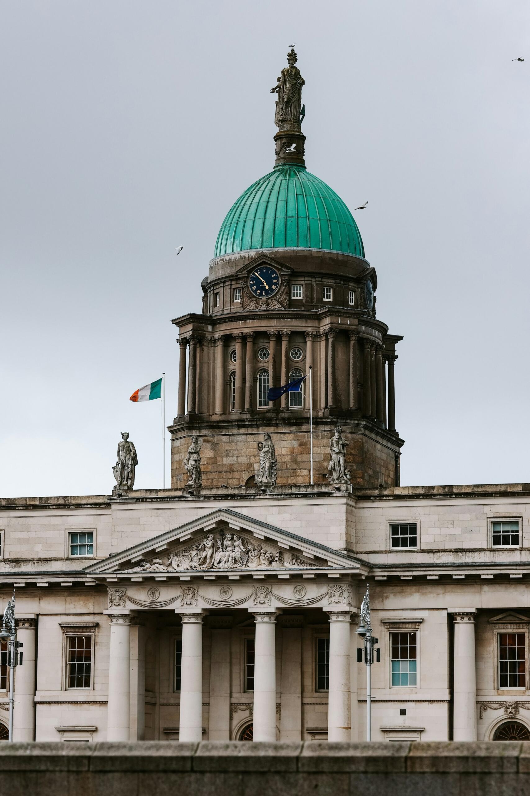 Close-up of a historic building with a green dome, clock, and statues, featuring the Irish flag against a cloudy sky.