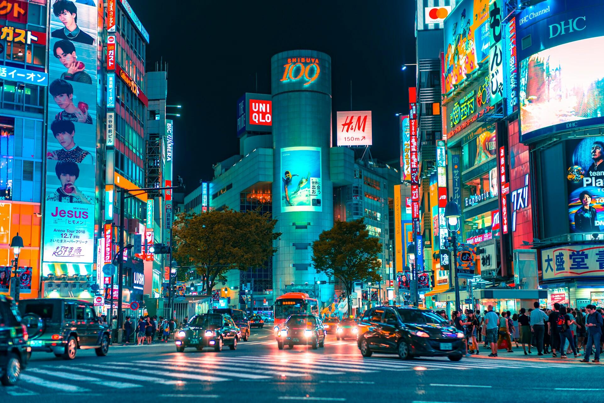 Vibrant neon lights illuminate a busy Shibuya intersection at night, featuring billboards and bustling traffic among pedestrians.